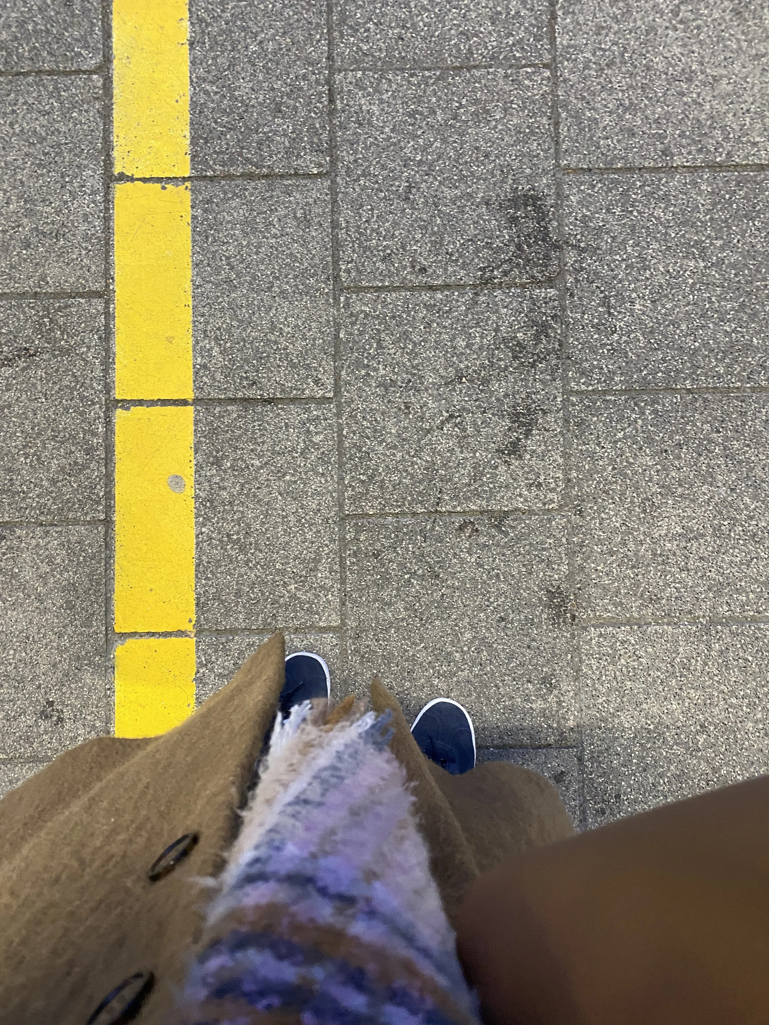 Overhead view of a person standing on a sidewalk near a yellow line, wearing navy shoes, a brown coat, and patterned pants.