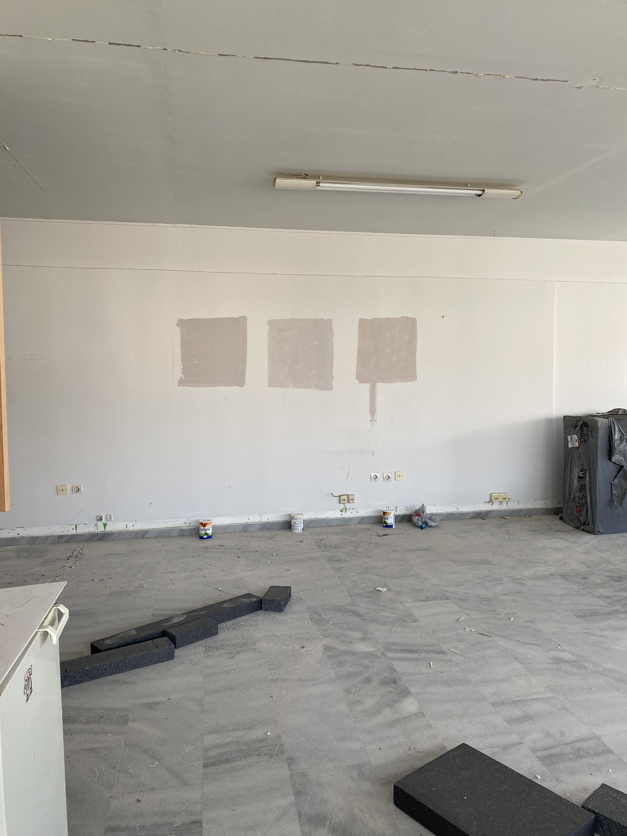 Room under renovation with three patches of primer on the white wall, construction materials on the floor, and an unfinished ceiling with a fluorescent light fixture.