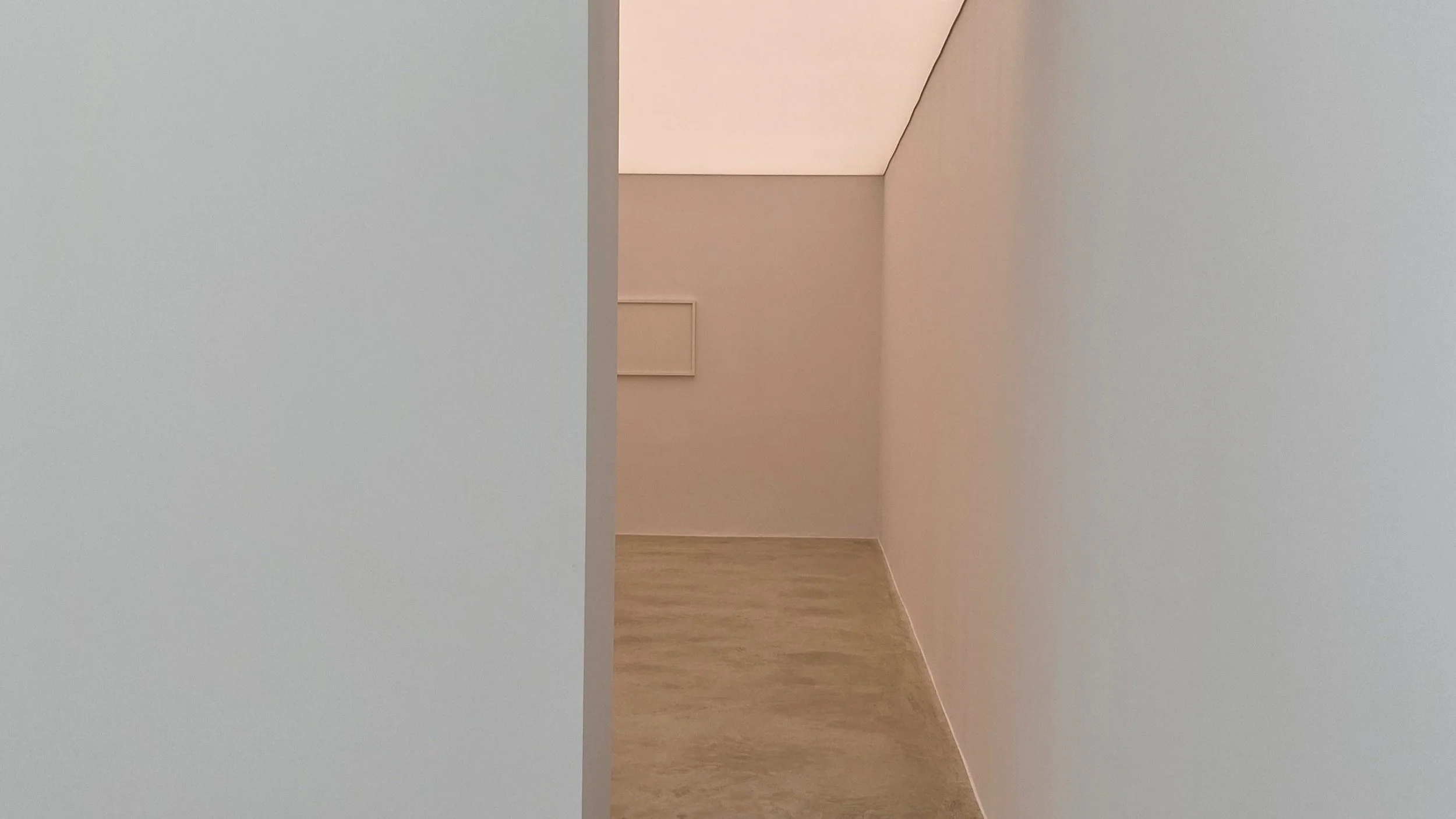 Minimalist interior view of a corner of a room with light-colored walls, wooden flooring, and a small framed picture on the far wall.