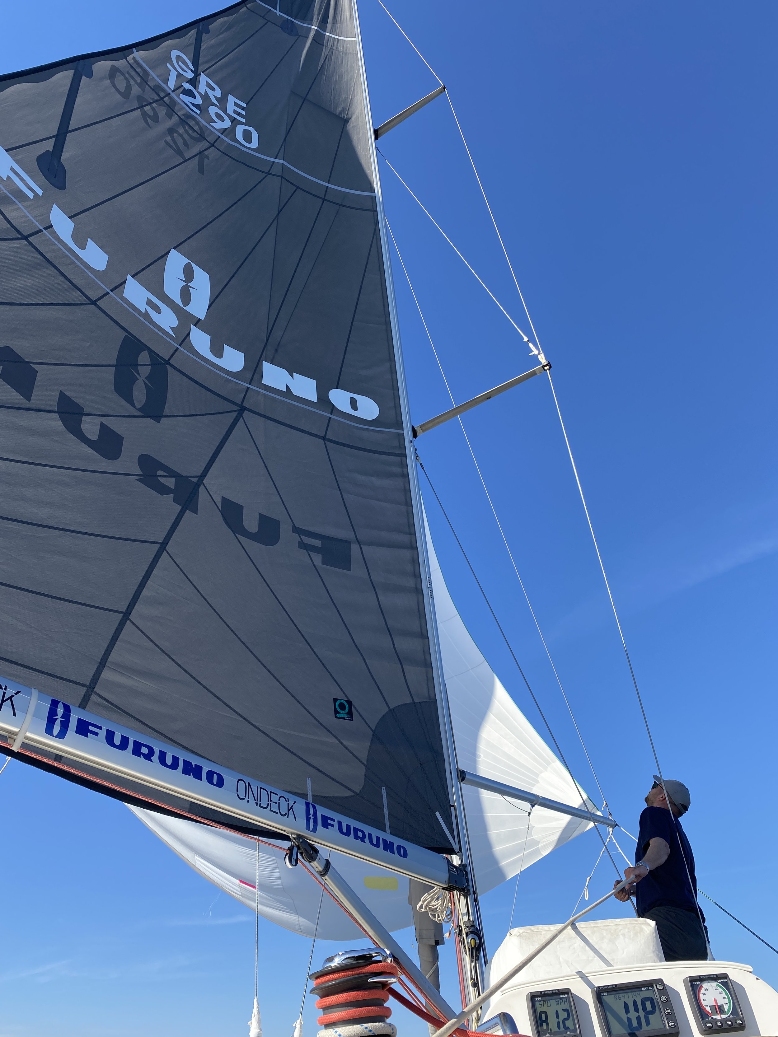 A person standing on a sailboat deck adjusting the sail, with clear blue sky in the background, and the sail displaying the words "FURUNO" and "ONDECK".
