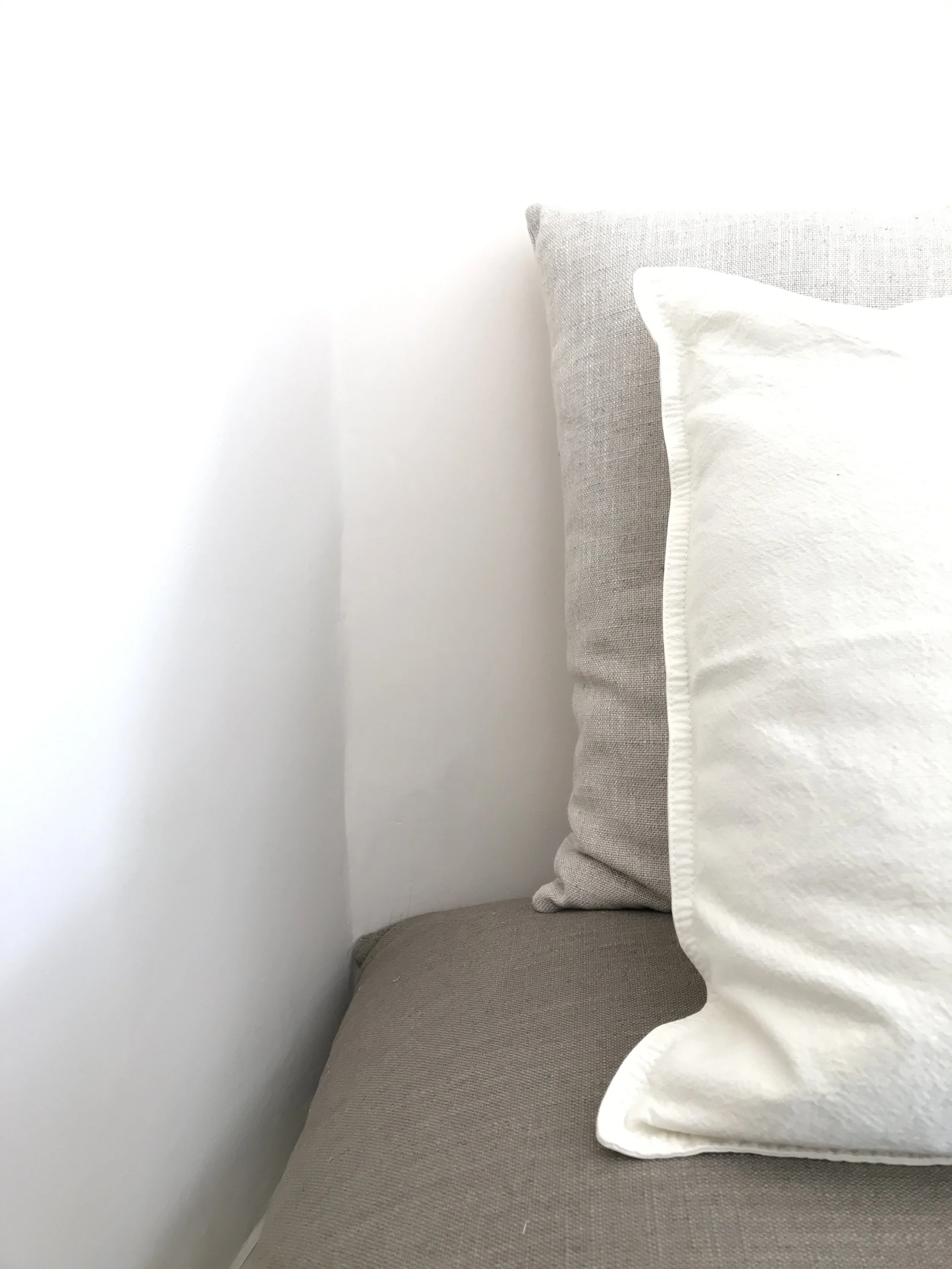 Close-up of a beige bed with a white pillow resting against a grey headboard, near a white wall.