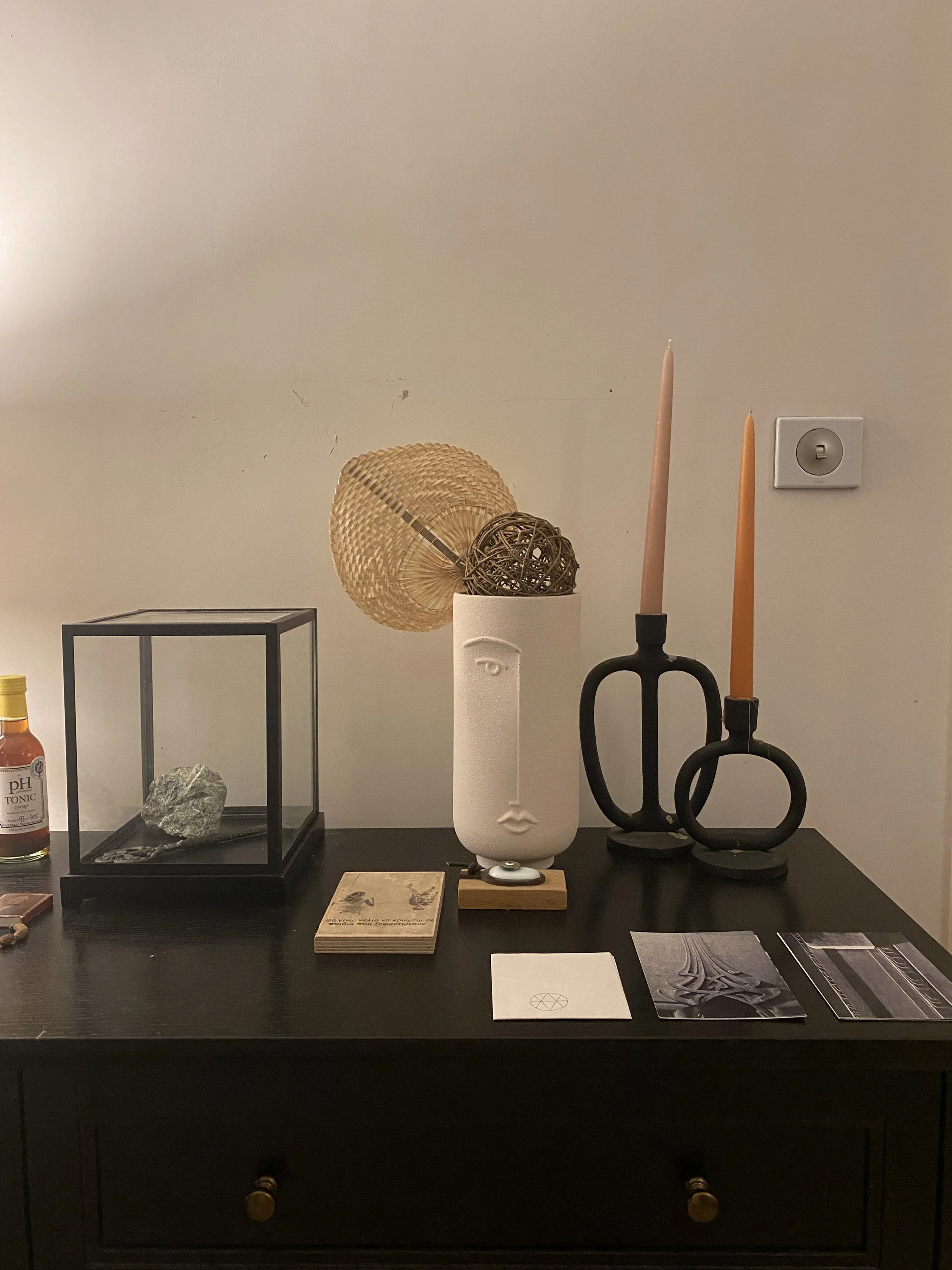 Decorative tabletop arrangement including a black geometric glass box with a rock and a small card inside, a white face-shaped sculpture with a wooden base holding a woven ball and straw fan, two tall candles in black holders with circular bases, and