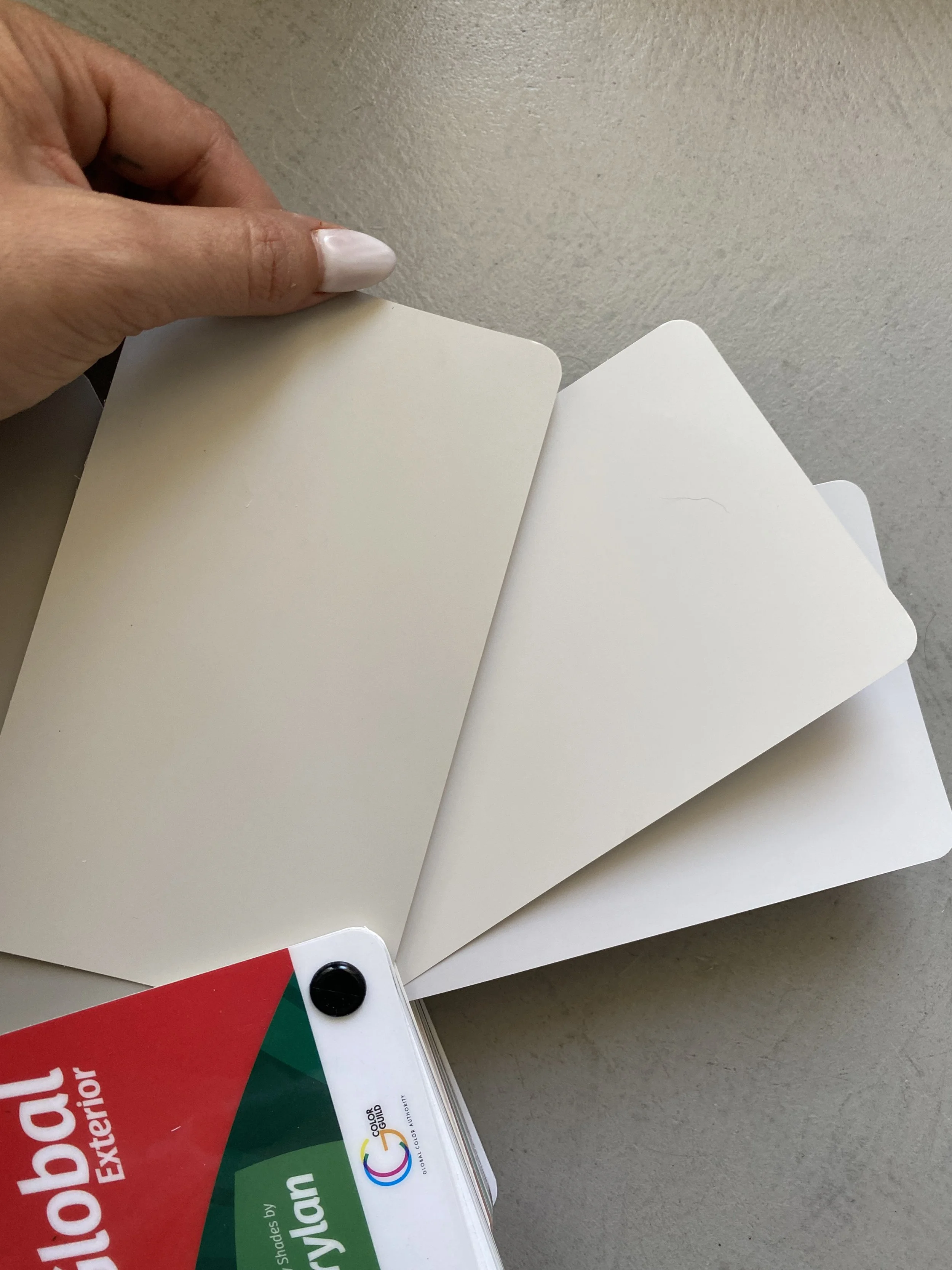 A hand with white painted nails flipping through multiple blank color sample cards in a fan shape, with a color swatch booklet at the bottom left corner.