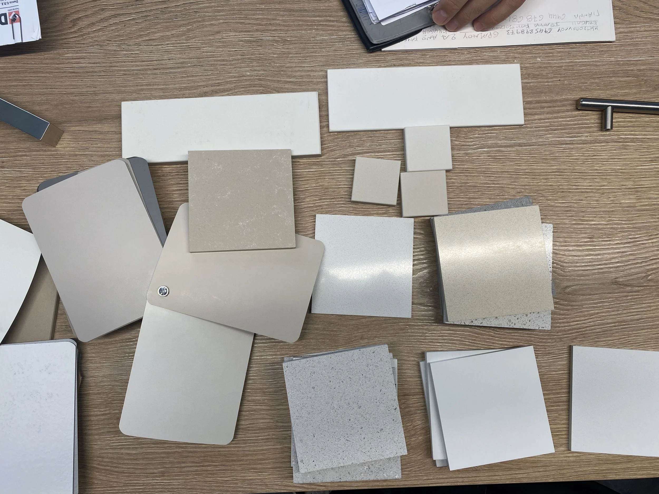 Various color swatches and sample tiles for interior design or construction arranged on a wooden table, with some notes and a person's hand visible at the top.