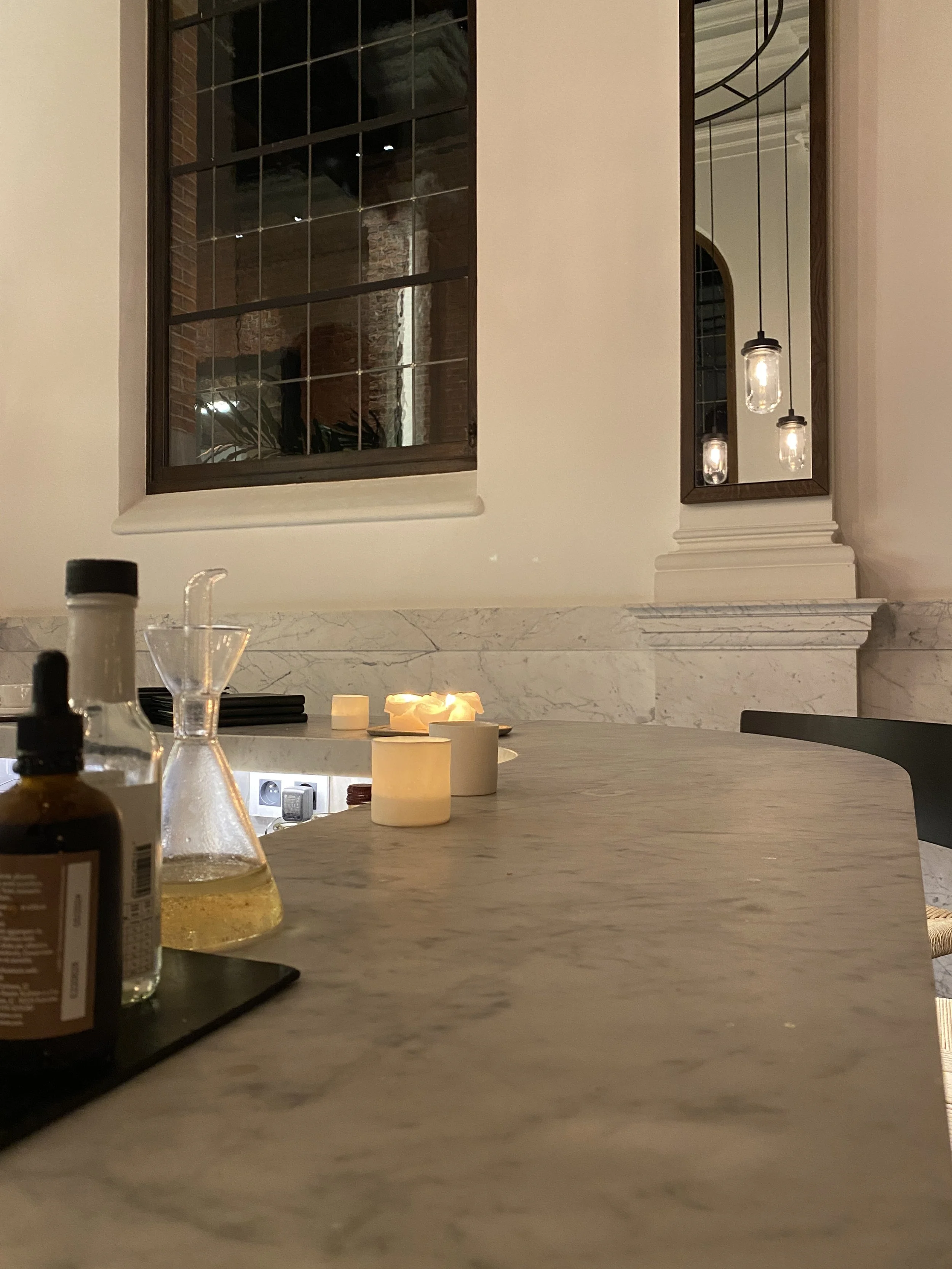 Close-up of a marble countertop with candles and bottles, interior of a room with large windows, hanging lights, and a mirror reflecting brick columns and industrial design elements.
