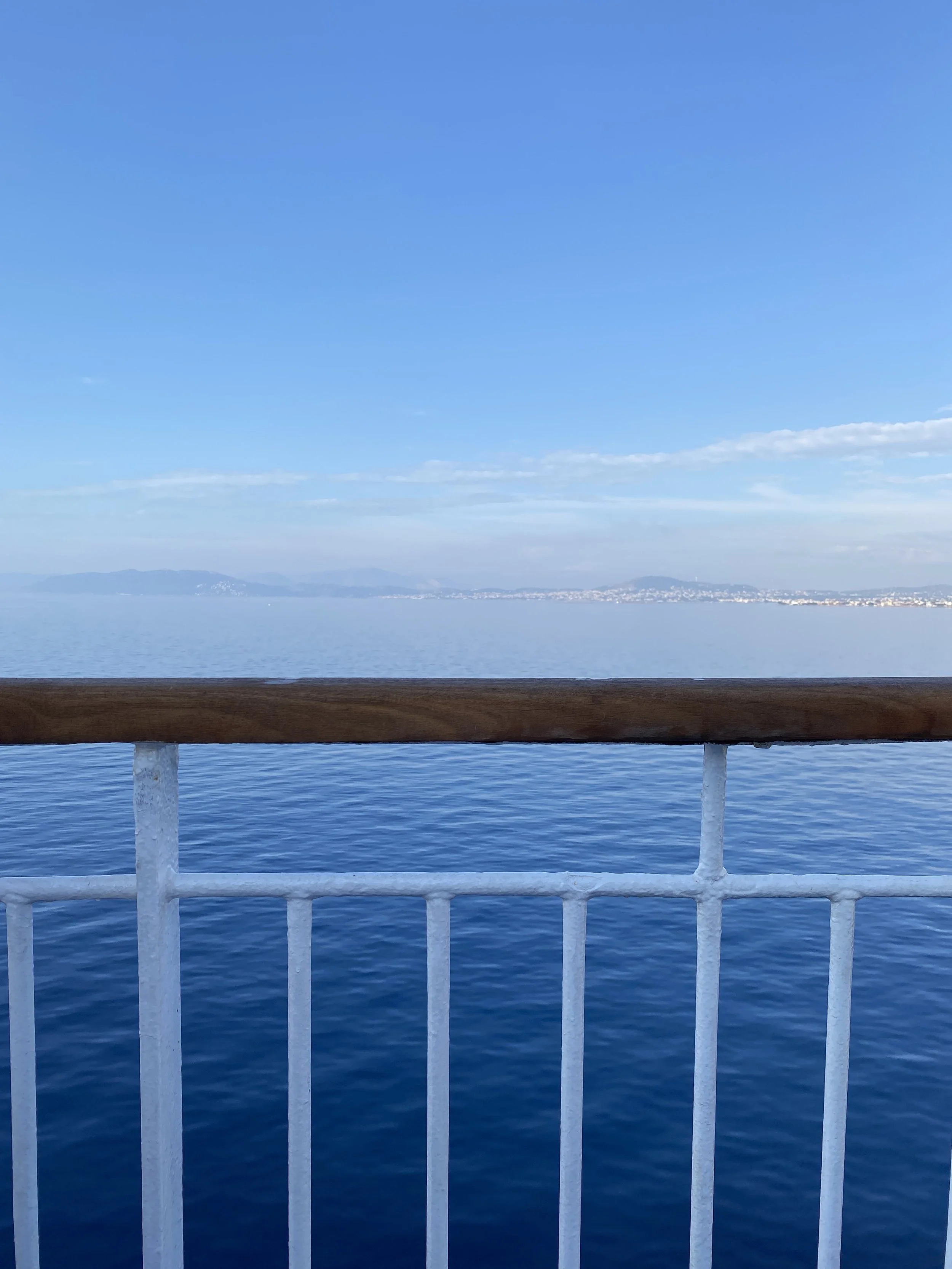 View from a boat or dock showing the water, distant mountains, and a coastline under a clear blue sky, with a wooden and white metal railing in the foreground.