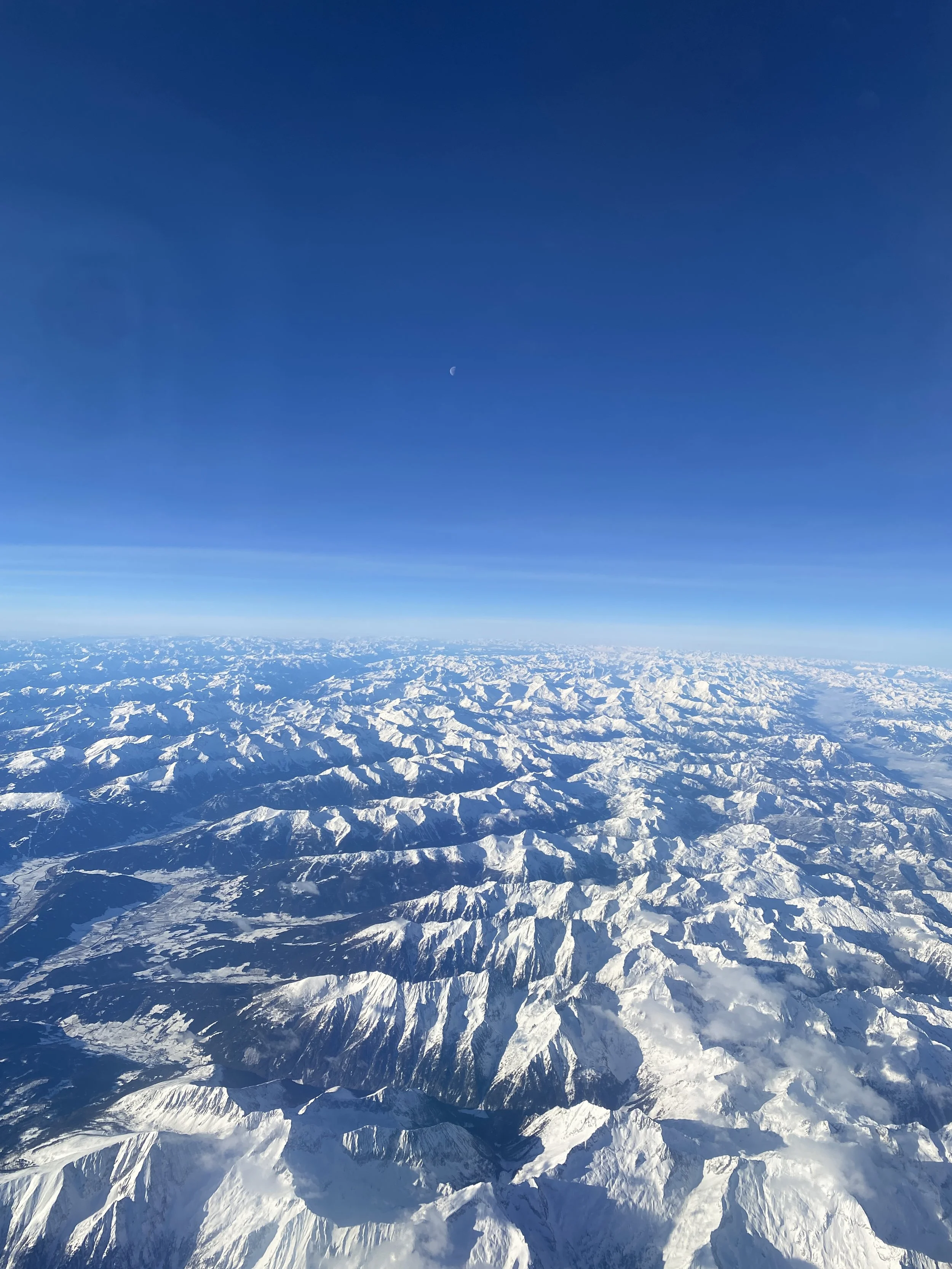 Aerial view of a snow-covered mountain range under a clear blue sky with the moon visible.