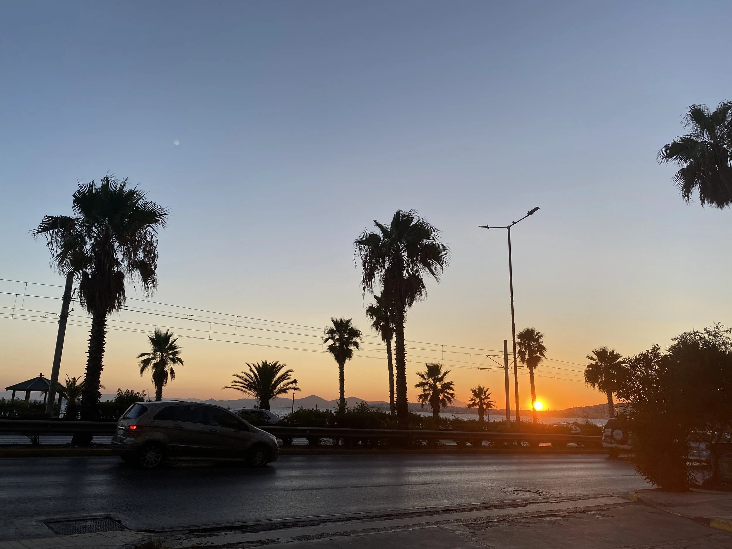 Sunset over a highway with palm trees, cars, power lines, a street lamp, and mountains in the distance.