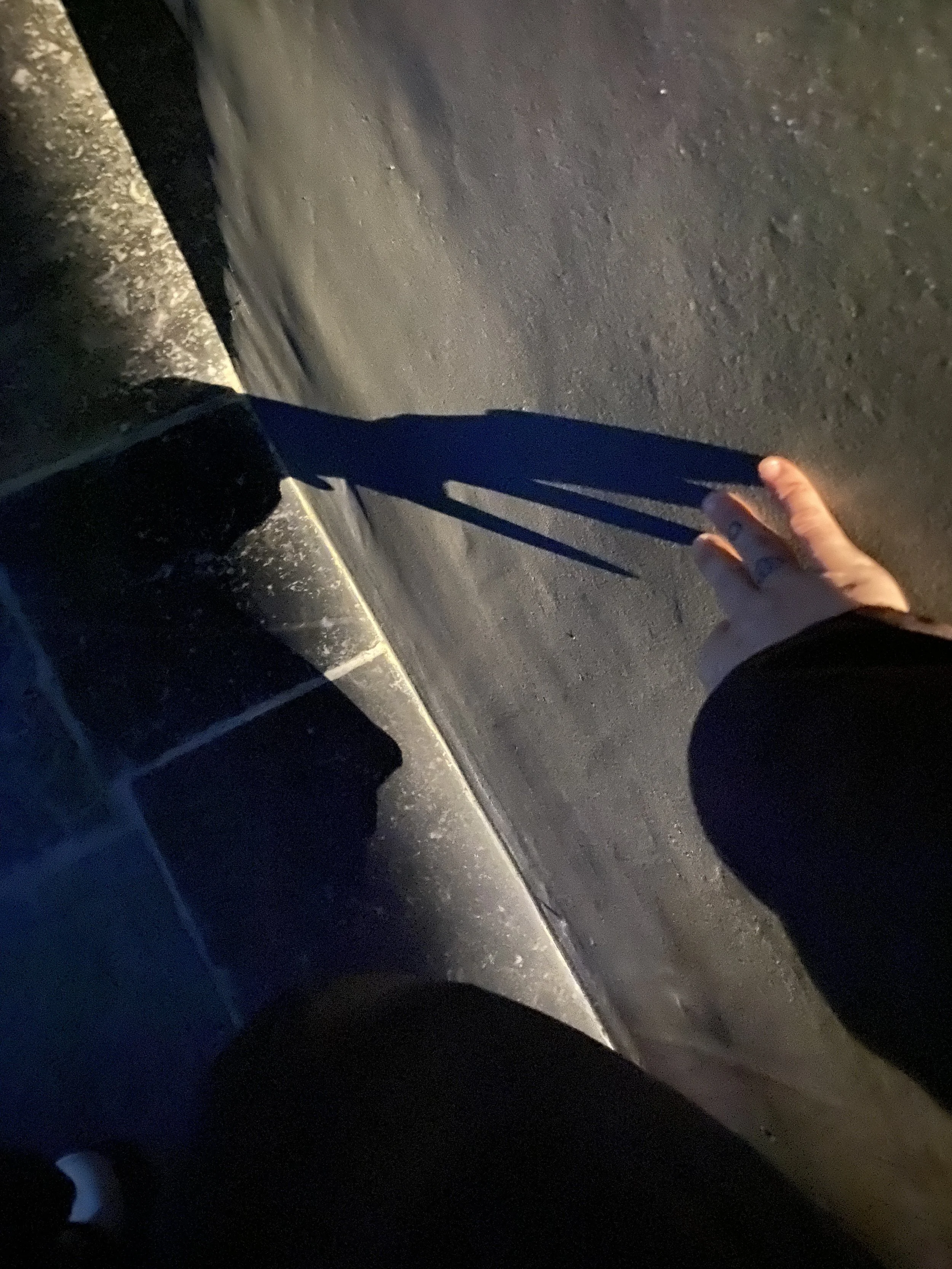 A person's hand with tattoos reaching out to touch the shadow of their hand cast on a road surface at night, with a vehicle wheel visible in the background.