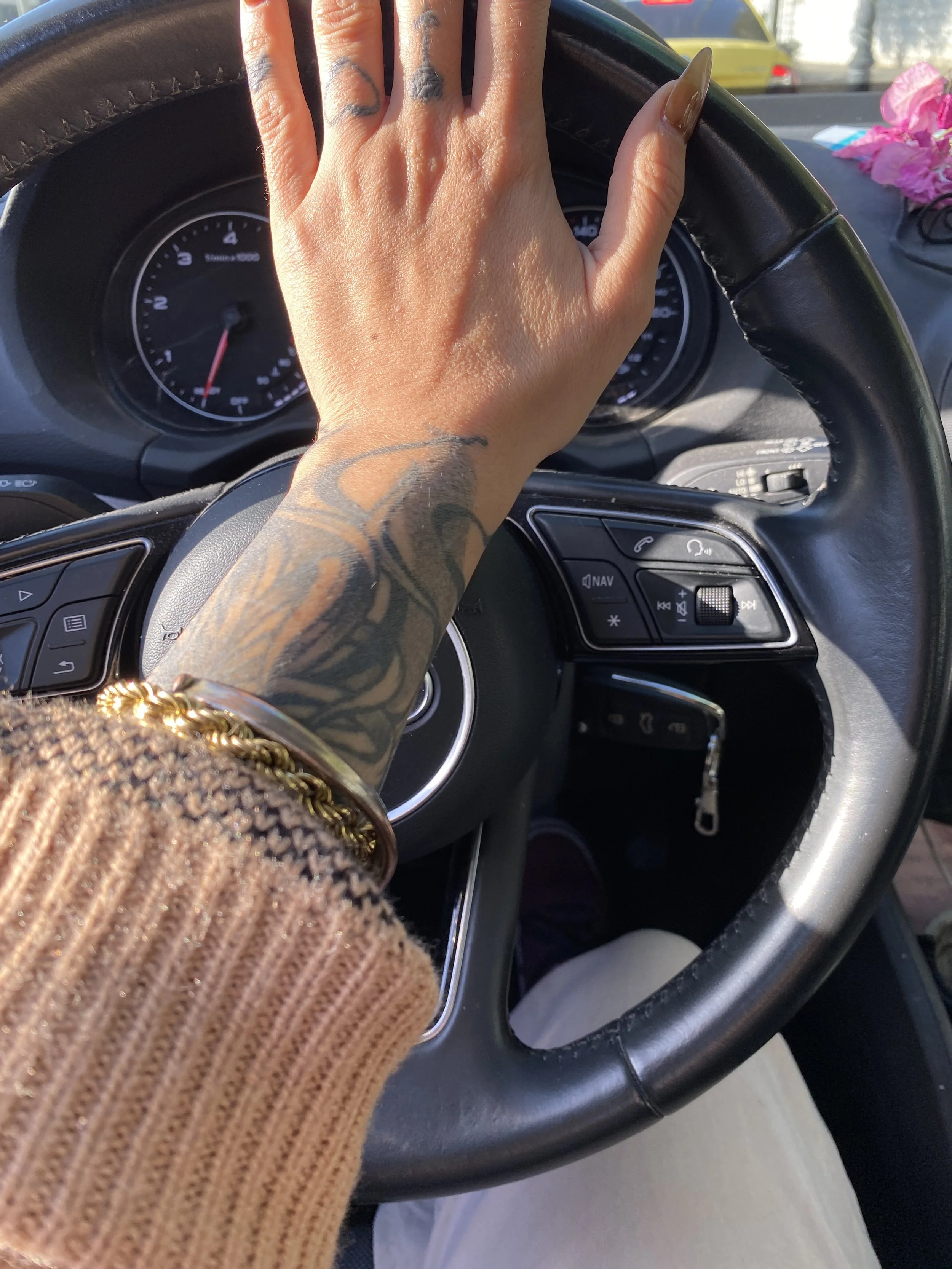 A person with tattooed hands and long nails resting their hand on a car's steering wheel while driving. The person is wearing a gold bracelet, and there are pink flowers and a yellow car visible in the background.