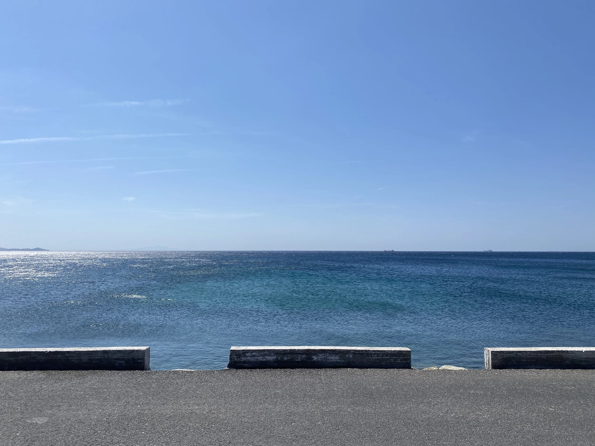 View of the ocean with a clear blue sky and some distant land or islands on the horizon, viewed from a pavement edge with concrete blocks in the foreground.
