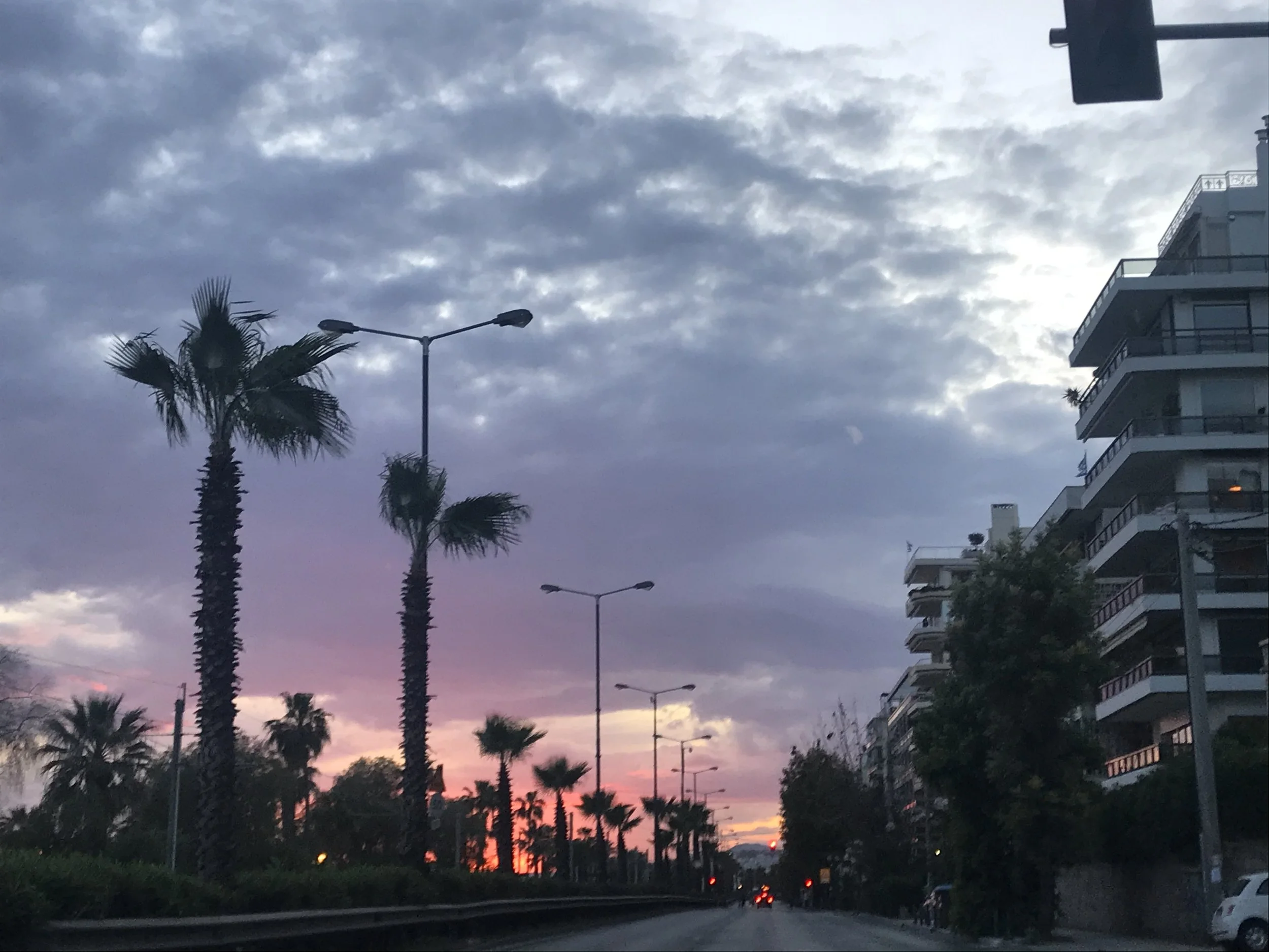 Skyscraper apartment buildings next to palm trees under a cloudy evening sky with a setting sun.