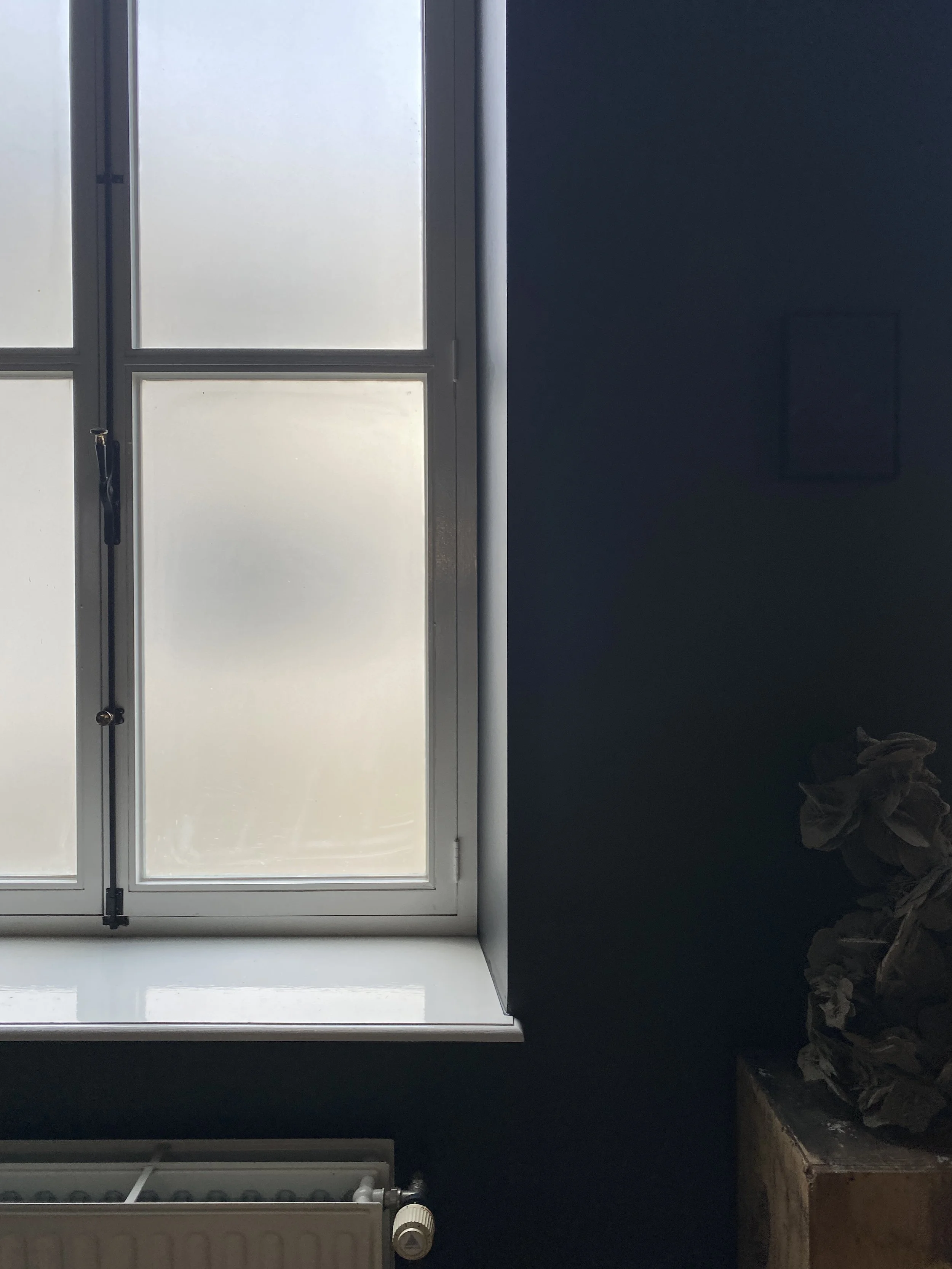 A frosted window with a white windowsill and a dark wall to the right, with some crumpled paper on a wooden surface.