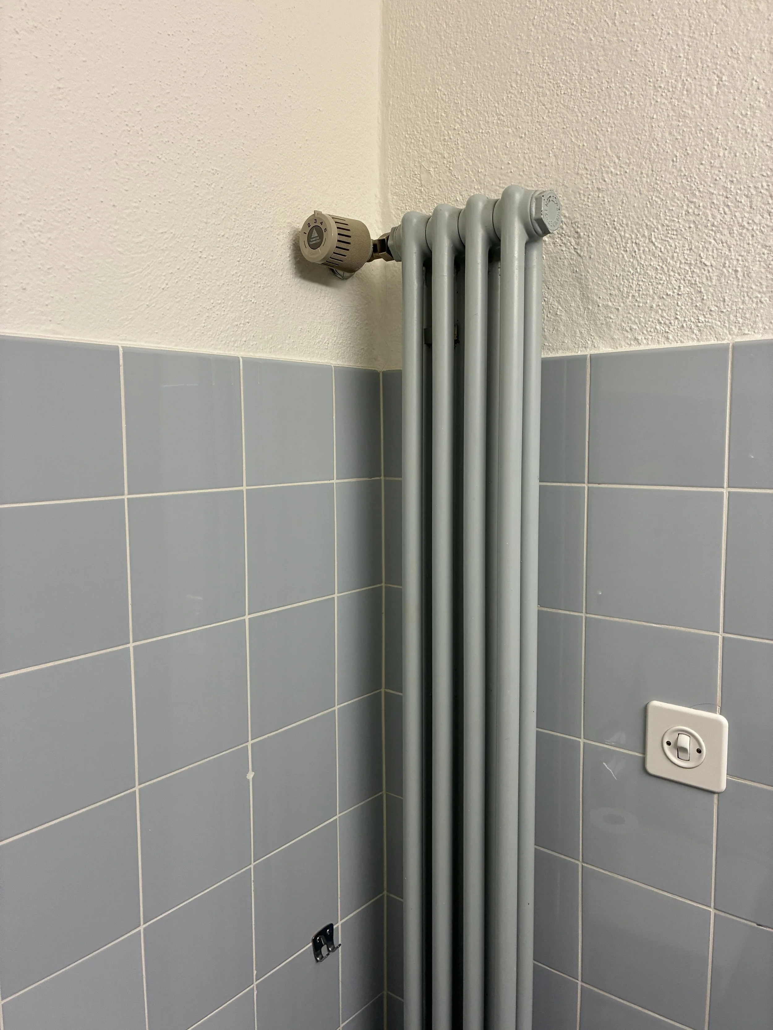 A corner of a room with light gray tiled walls and an electric radiator with four vertical pipes and a beige thermostat attached to the top left pipe.