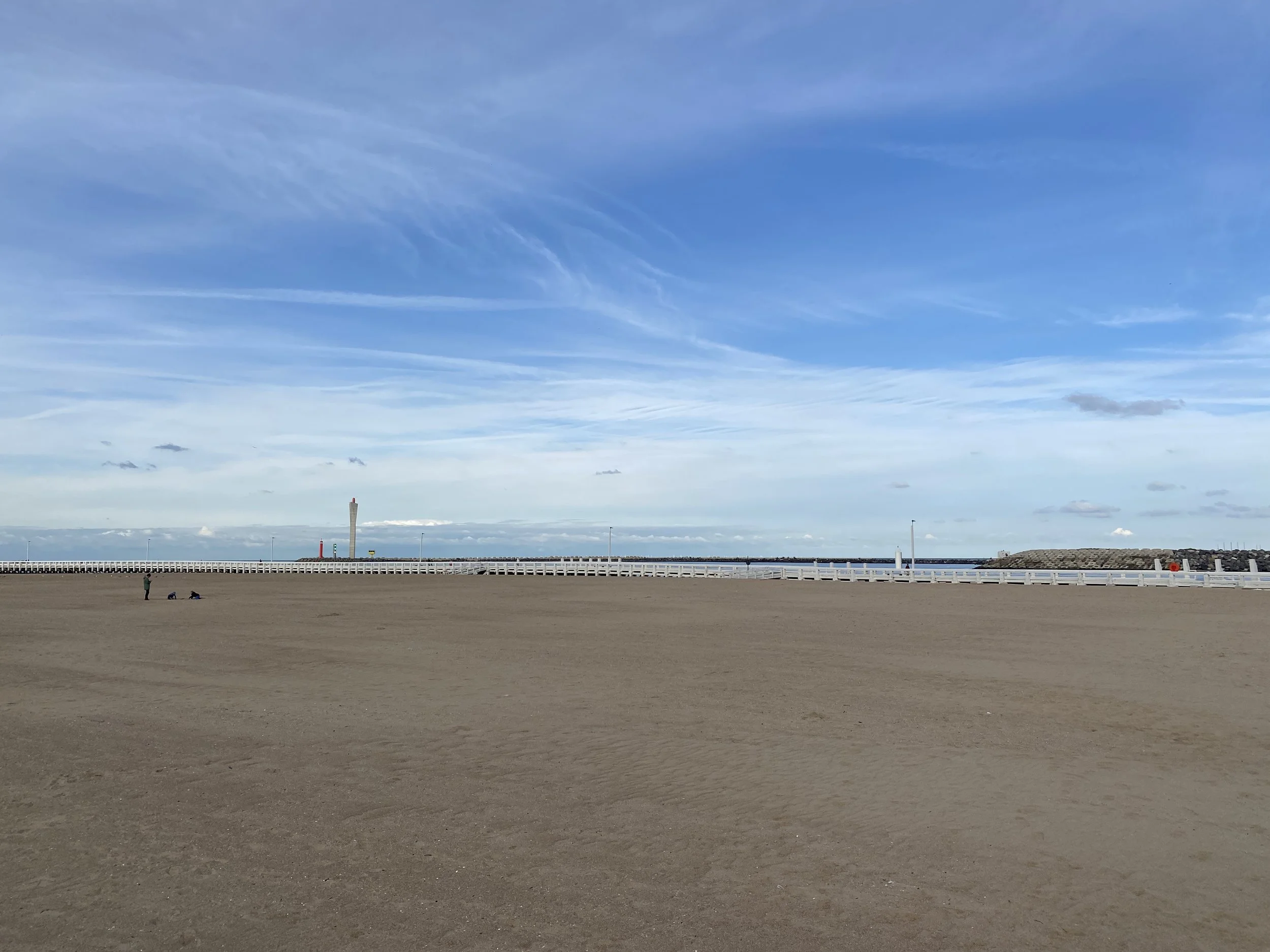 Empty sandy beach with a person and a dog, white railing, lighthouse and breakwater in the distance, partly cloudy sky with blue tones.