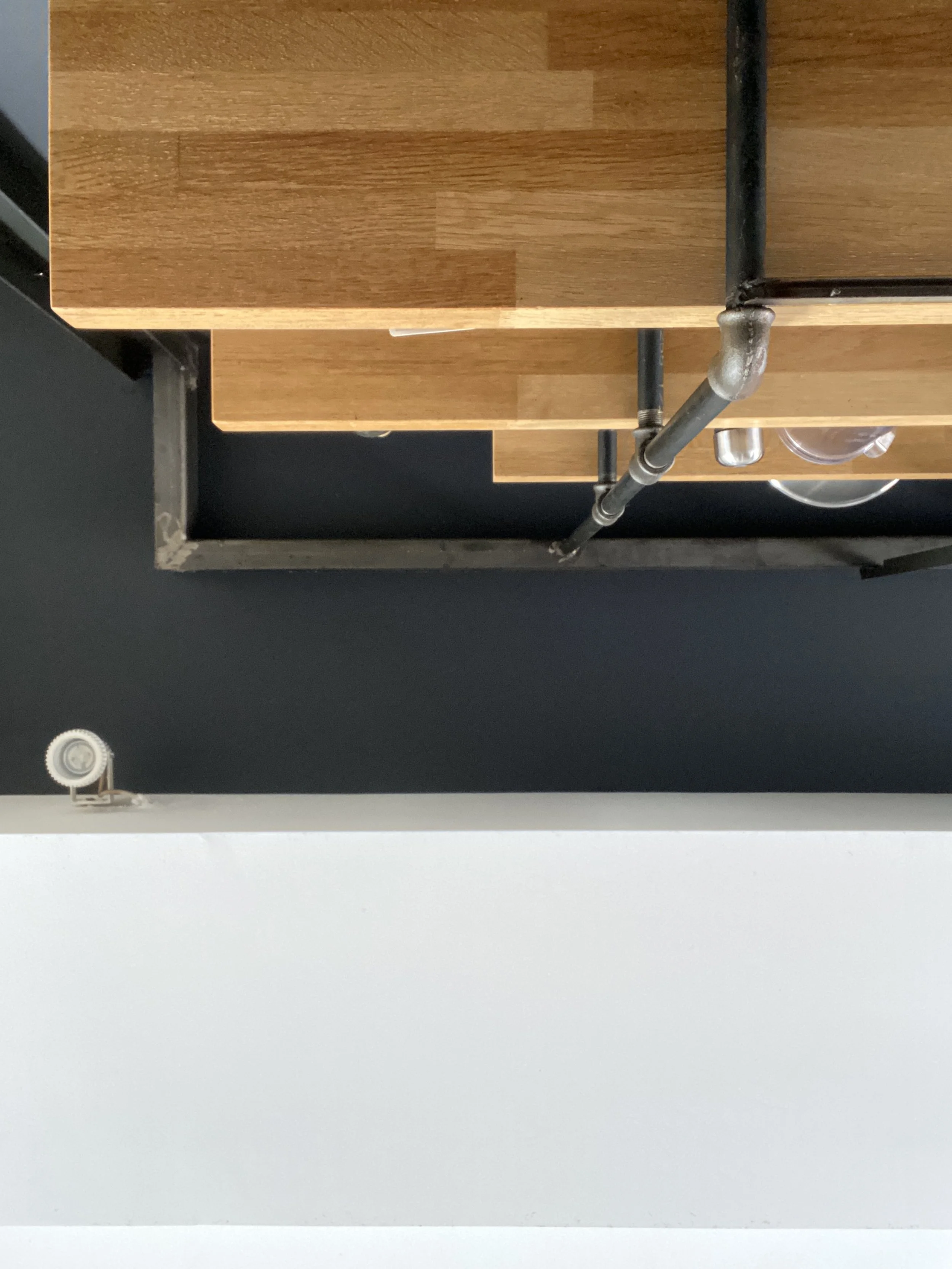 Top-down view of a wooden countertop with black piping beneath it, against a dark wall, and a white wall in the foreground.