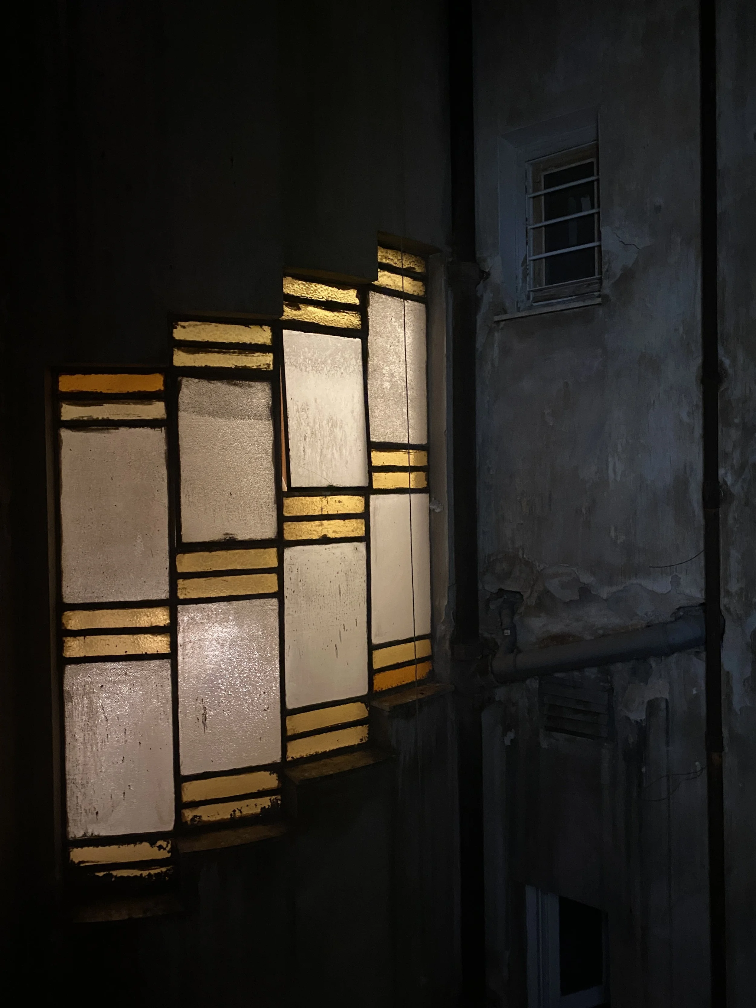 Dimly lit room with old, cracked walls, an uneven window with flush bars, and a large rectangular stained glass window with yellow and frosted glass panes, some of which are broken or missing.