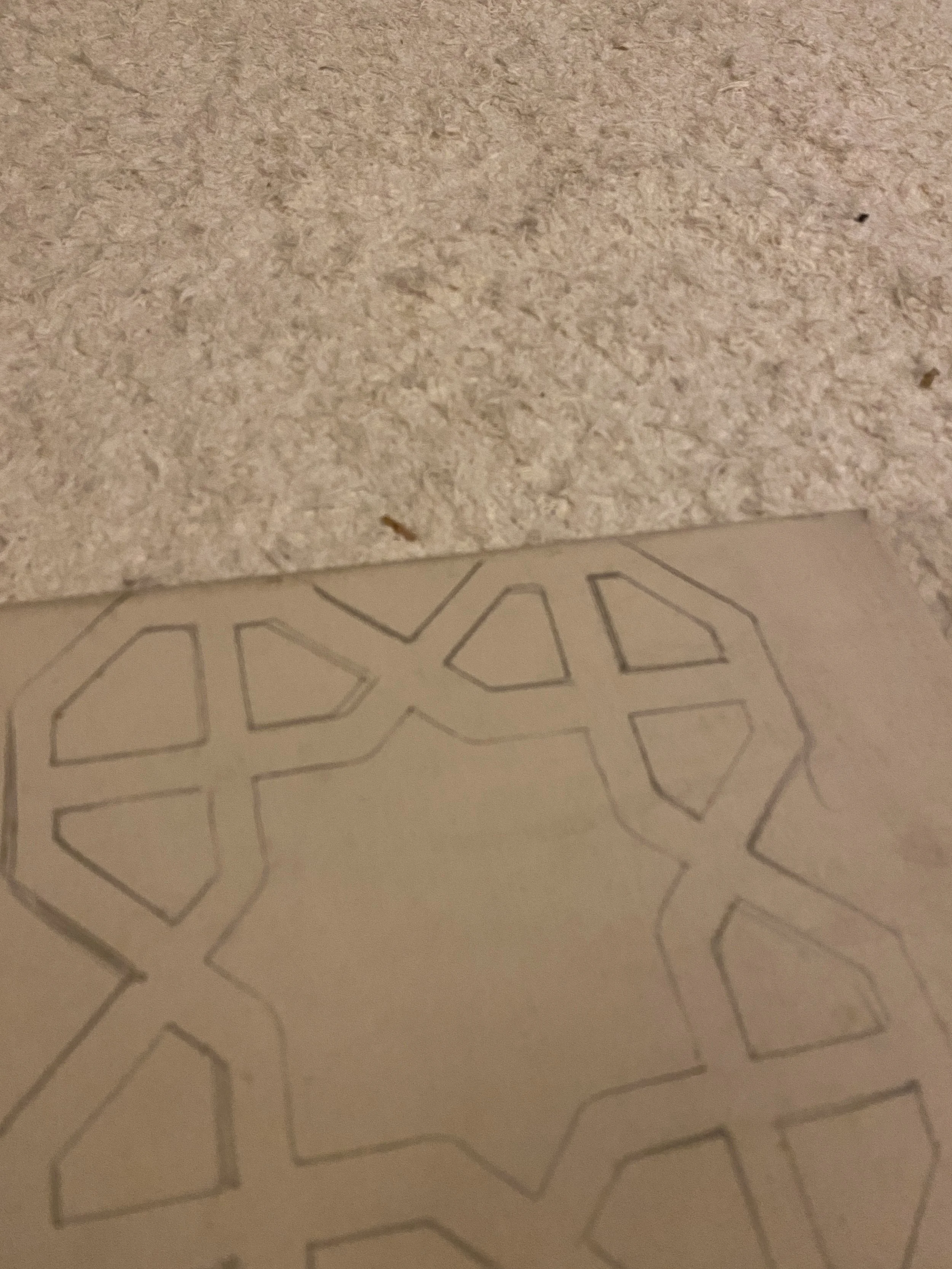 A piece of paper with a hand-drawn geometric star pattern in pencil, placed on a textured beige carpet.