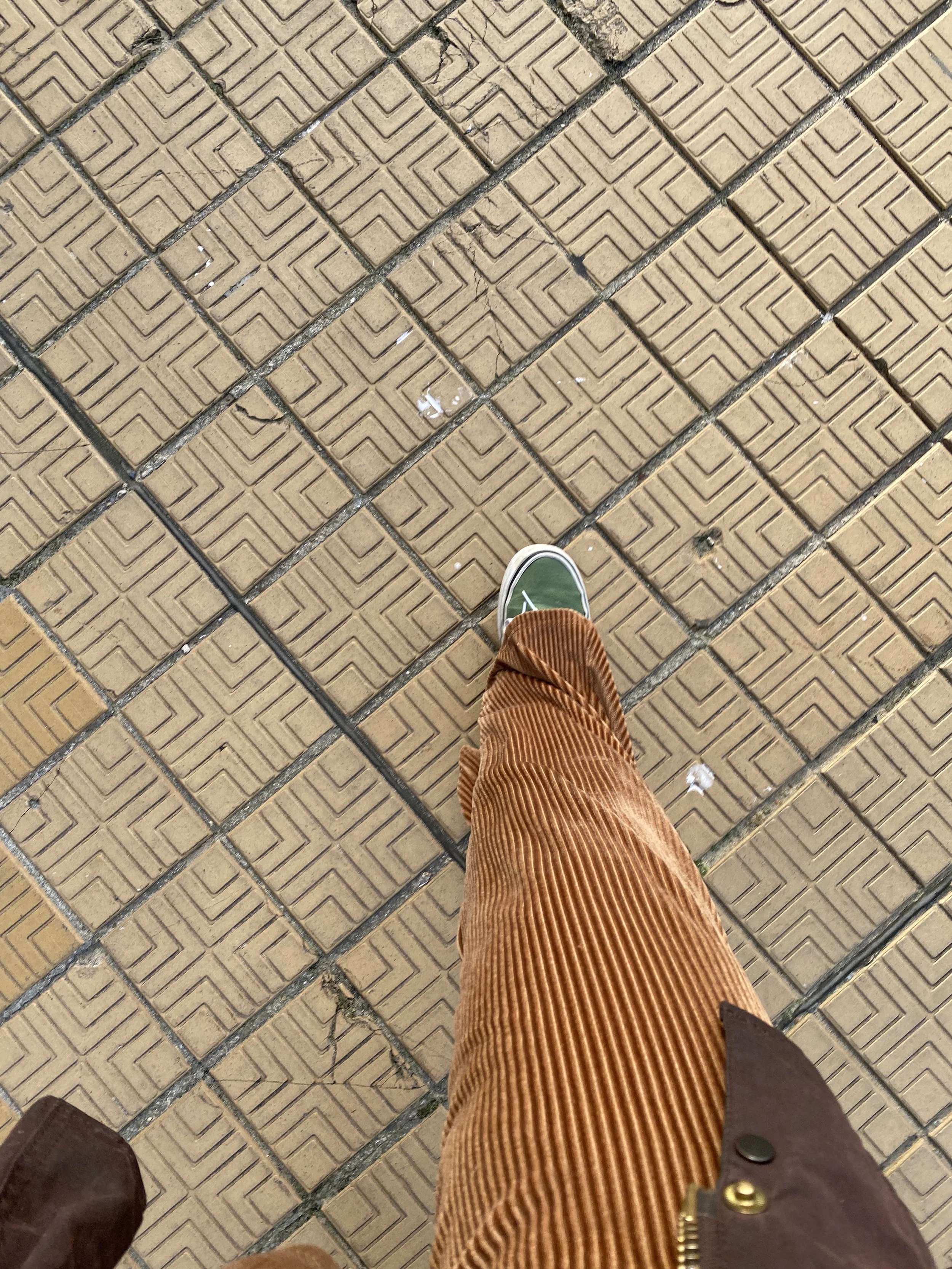 Top-down view of a person's leg and foot, wearing a green sneaker with white sole, brown corduroy pants, and a brown jacket, walking on a tiled sidewalk with a geometric pattern.