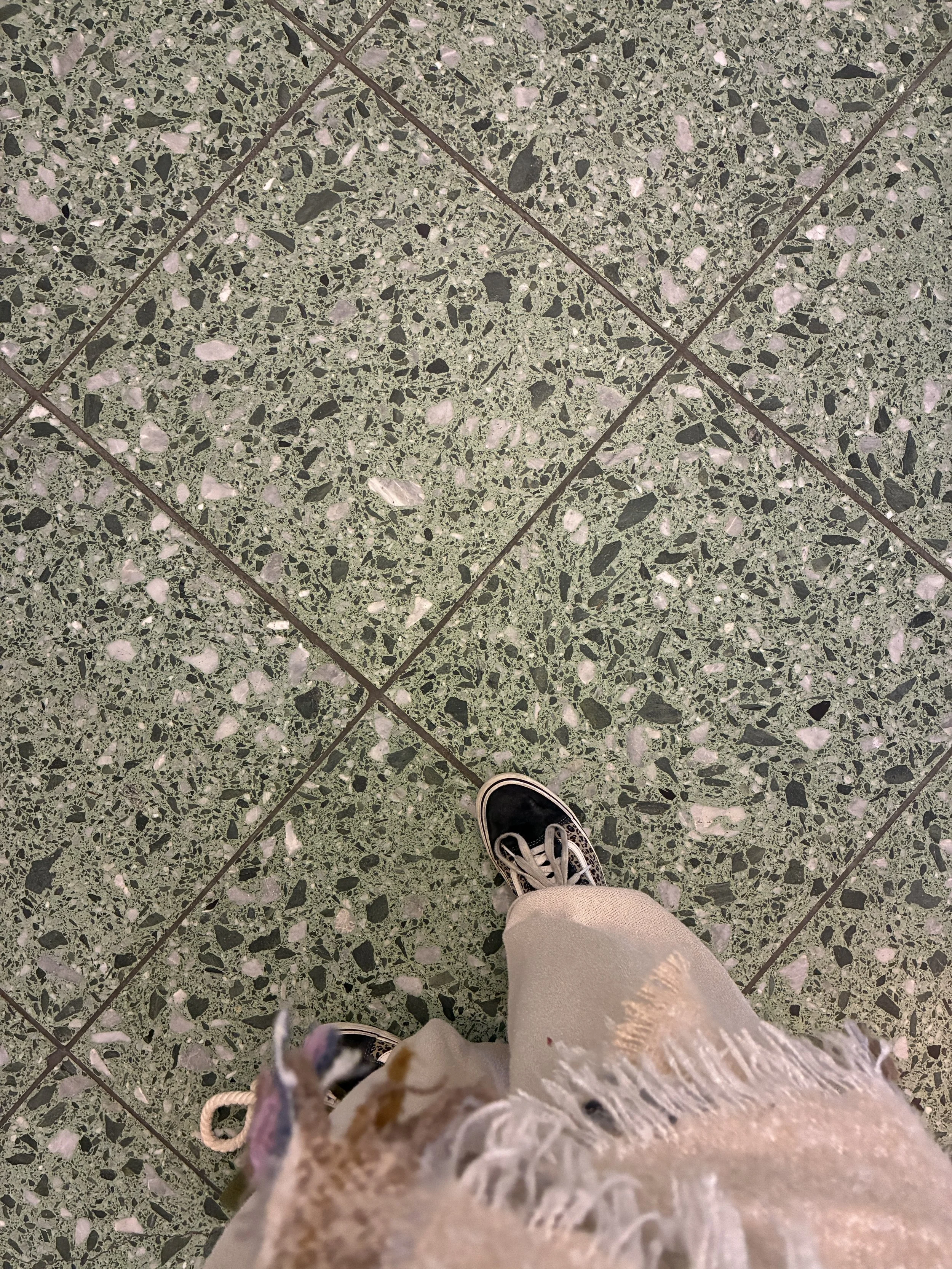 View of terrazzo floor tiles with a person's feet wearing black slip-on shoes and beige pants, with frayed edges on the pants.