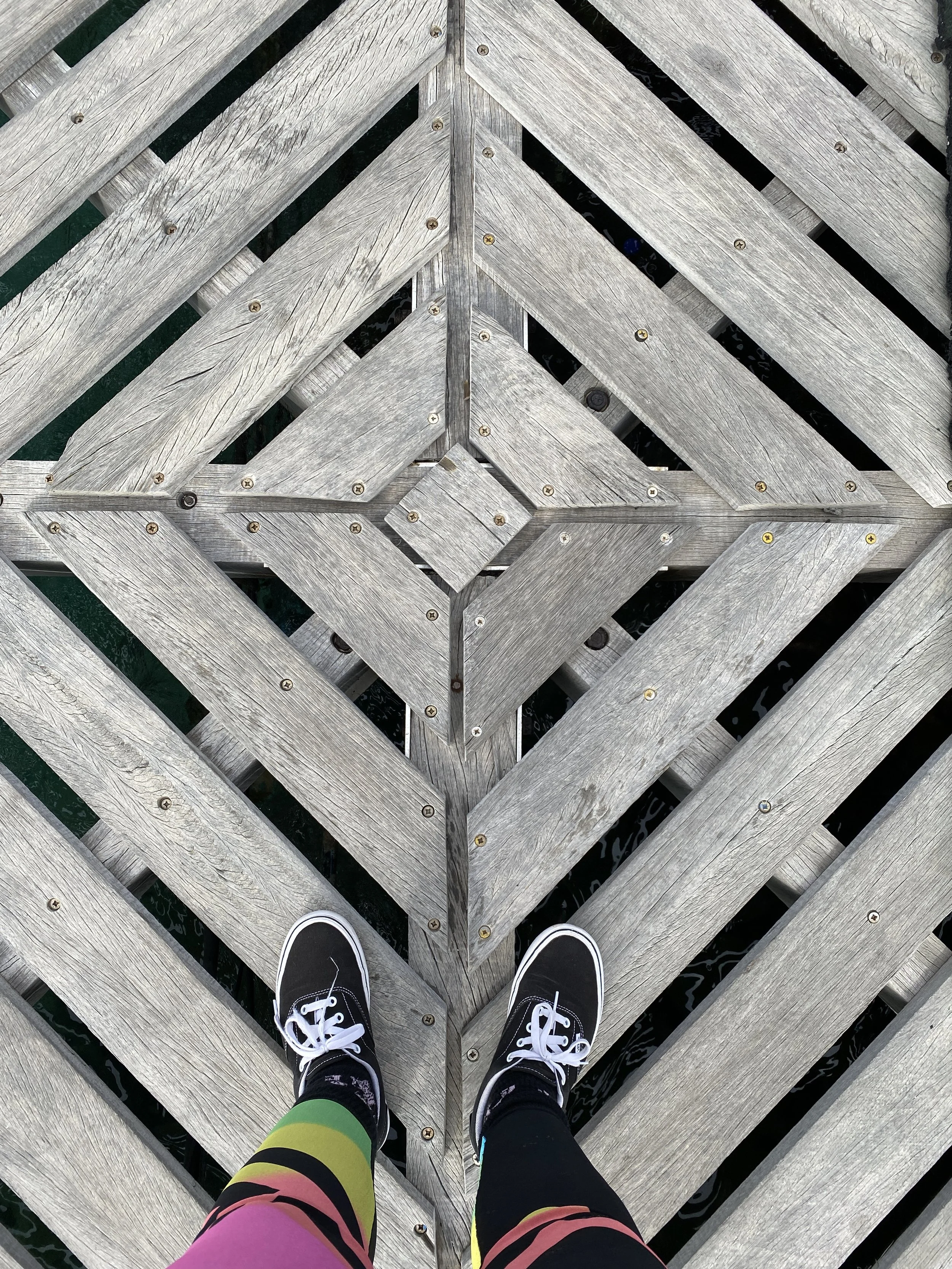 A person's feet wearing black sneakers with white laces and colorful striped leggings, standing on a weathered wooden surface with a diamond-shaped pattern.