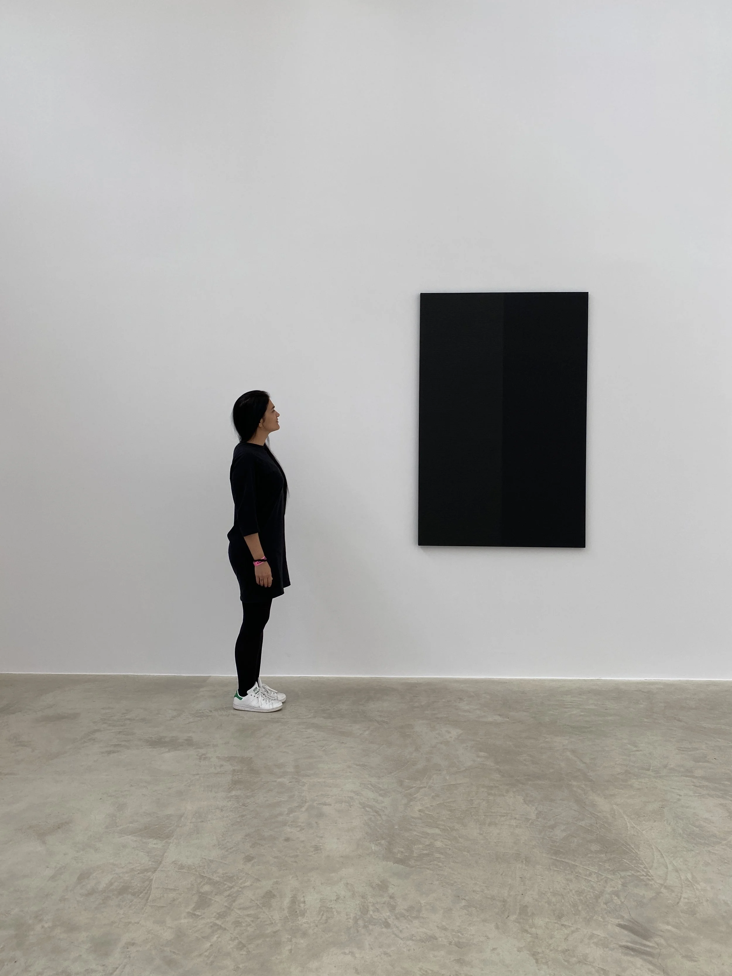 A woman stands in an art gallery looking at a large, black rectangular artwork on the white wall.