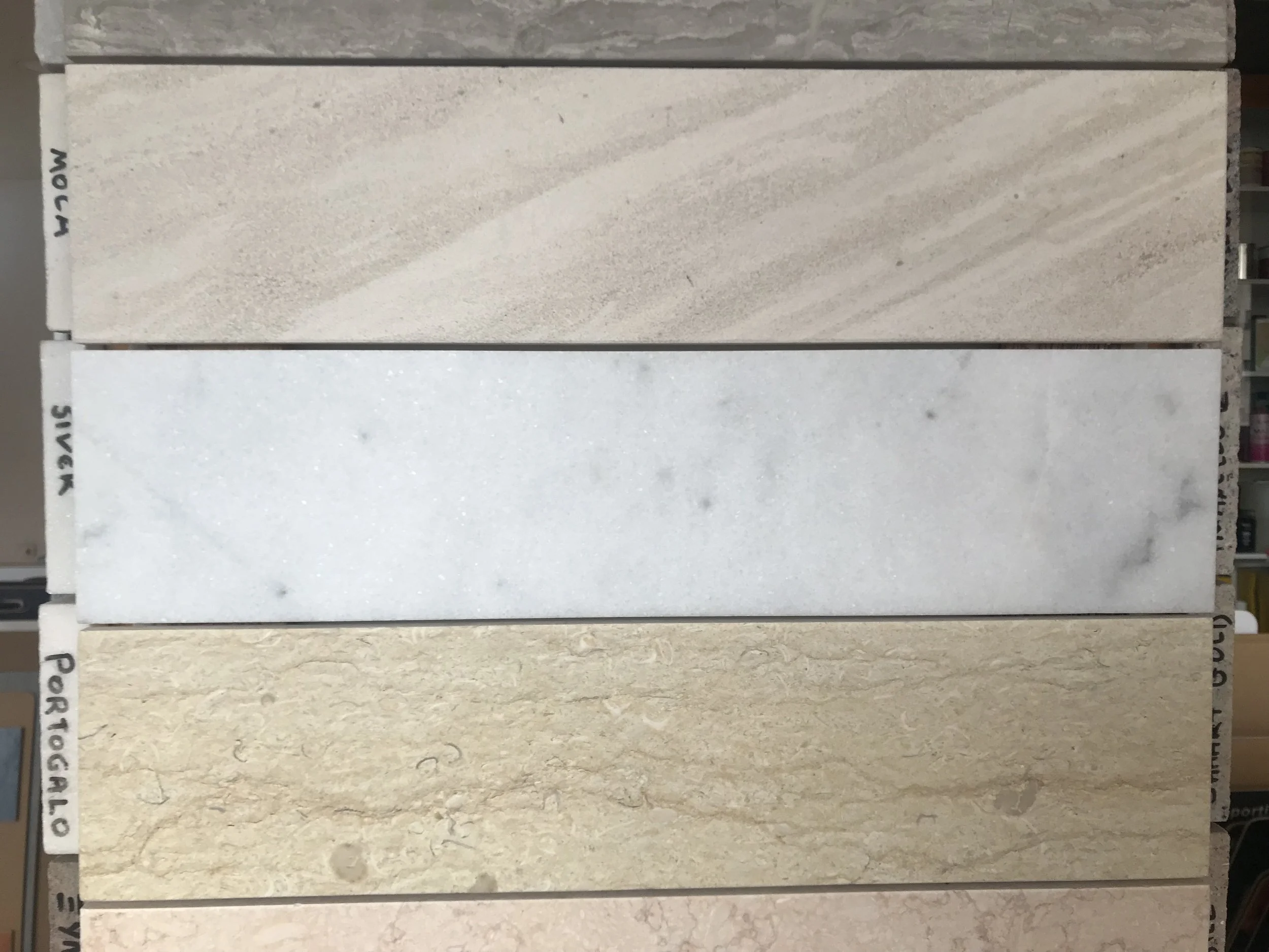 A display of five large rectangular sample tiles in beige and cream tones with different textures and patterns, arranged vertically on a tile store shelf.
