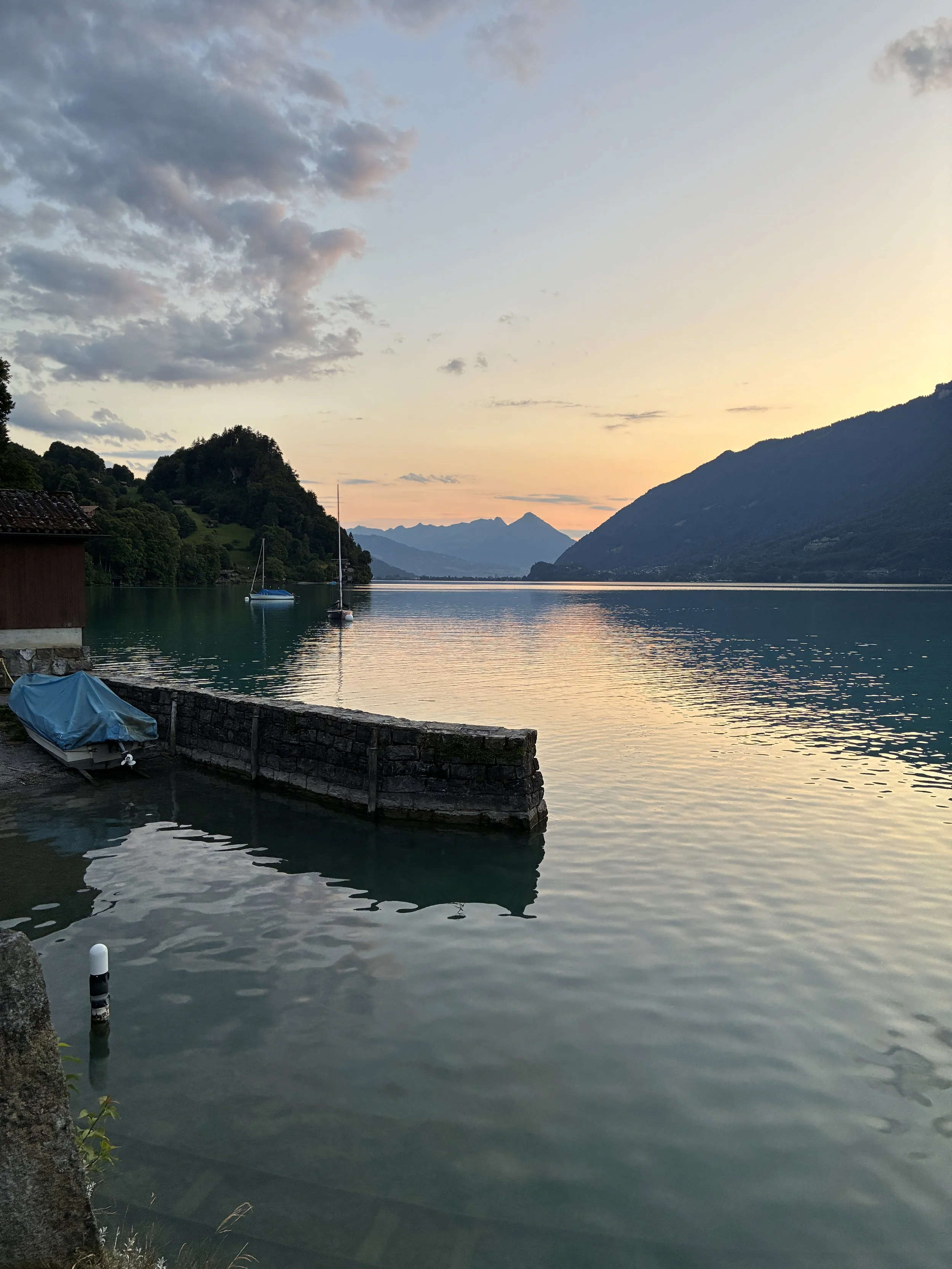 A peaceful lake at sunset with sailboats, mountains in the background, and a small dock with a covered boat in the foreground.