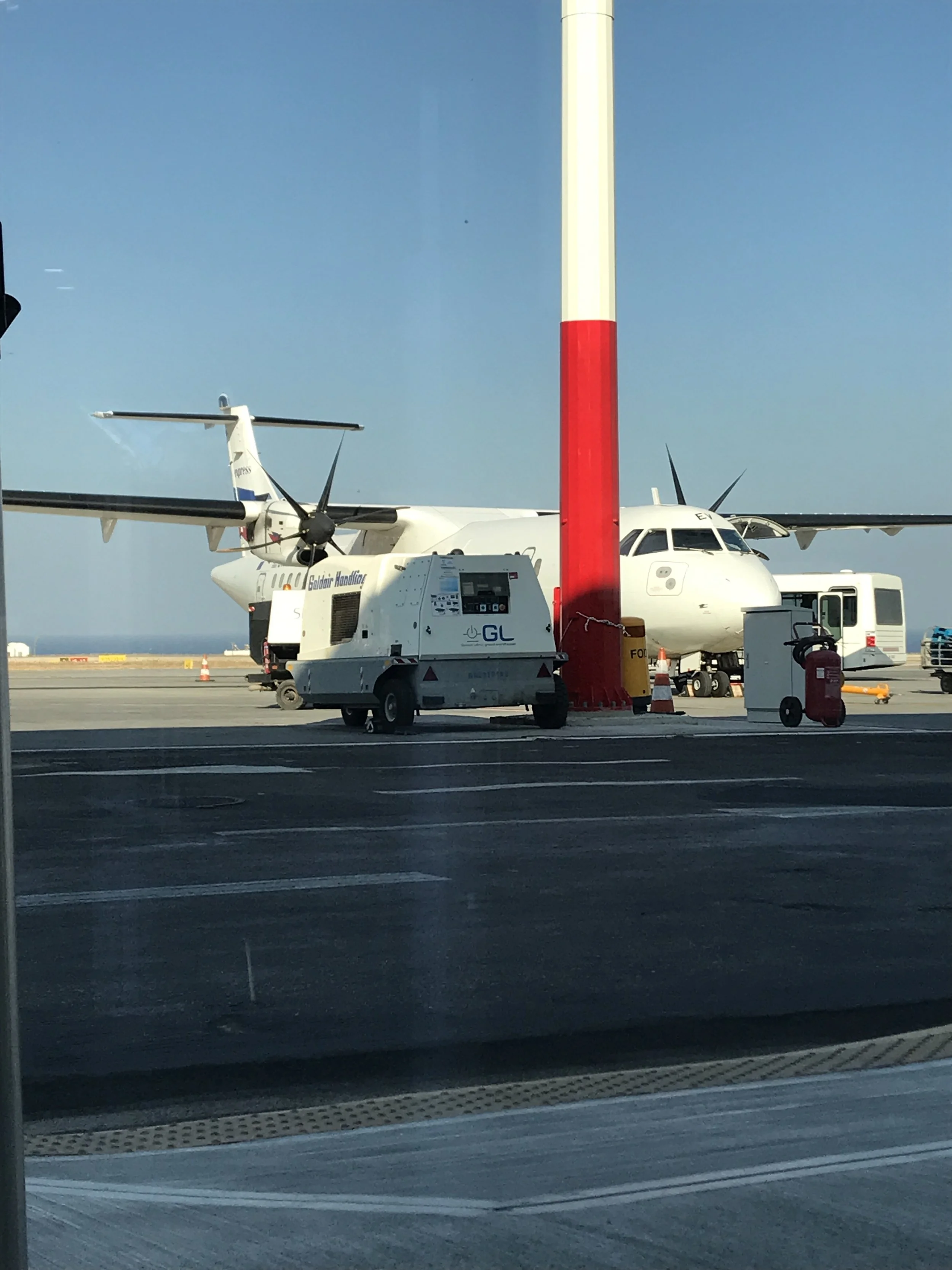 Inside an airport terminal behind a glass window, a small private jet is parked on the tarmac, with ground support equipment nearby, including a fuel truck, an orange safety cone, and other machinery, under a clear blue sky.