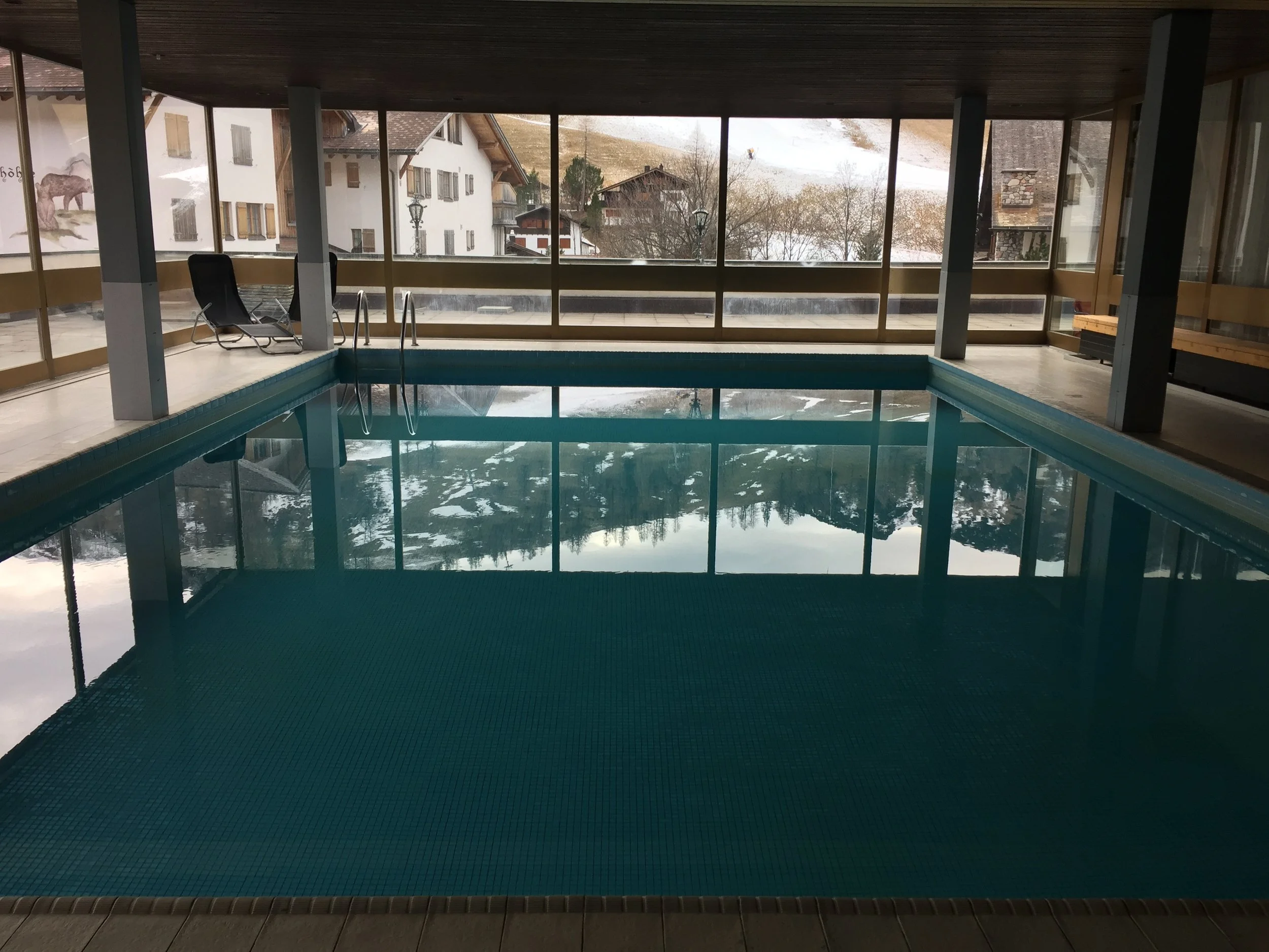 Indoor swimming pool with glass windows showing snow-covered houses and mountains outside, lounge chairs beside the pool.
