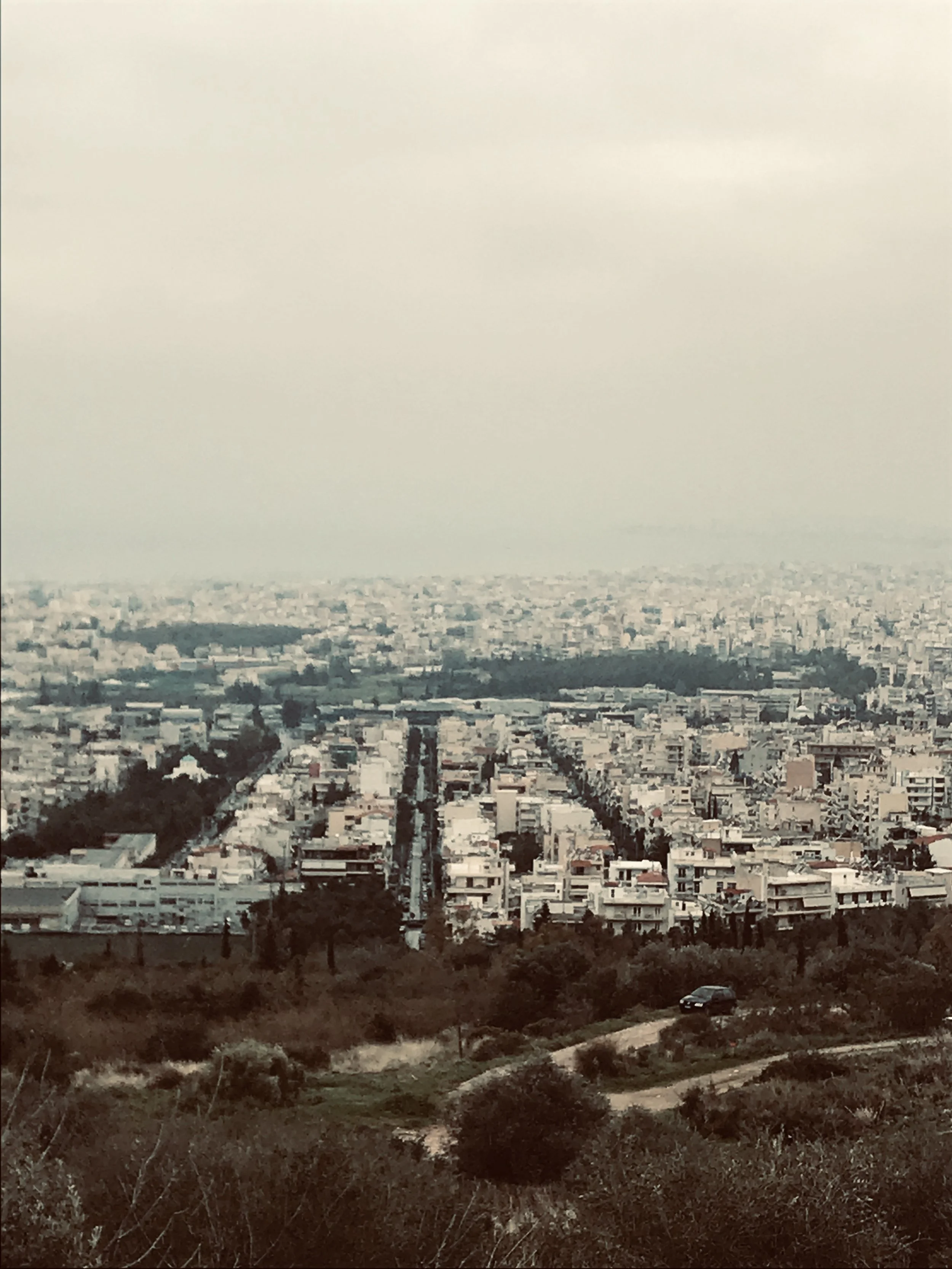 A cityscape with numerous white buildings seen from a hillside. The foreground shows dry, brownish vegetation and a road with a black car.