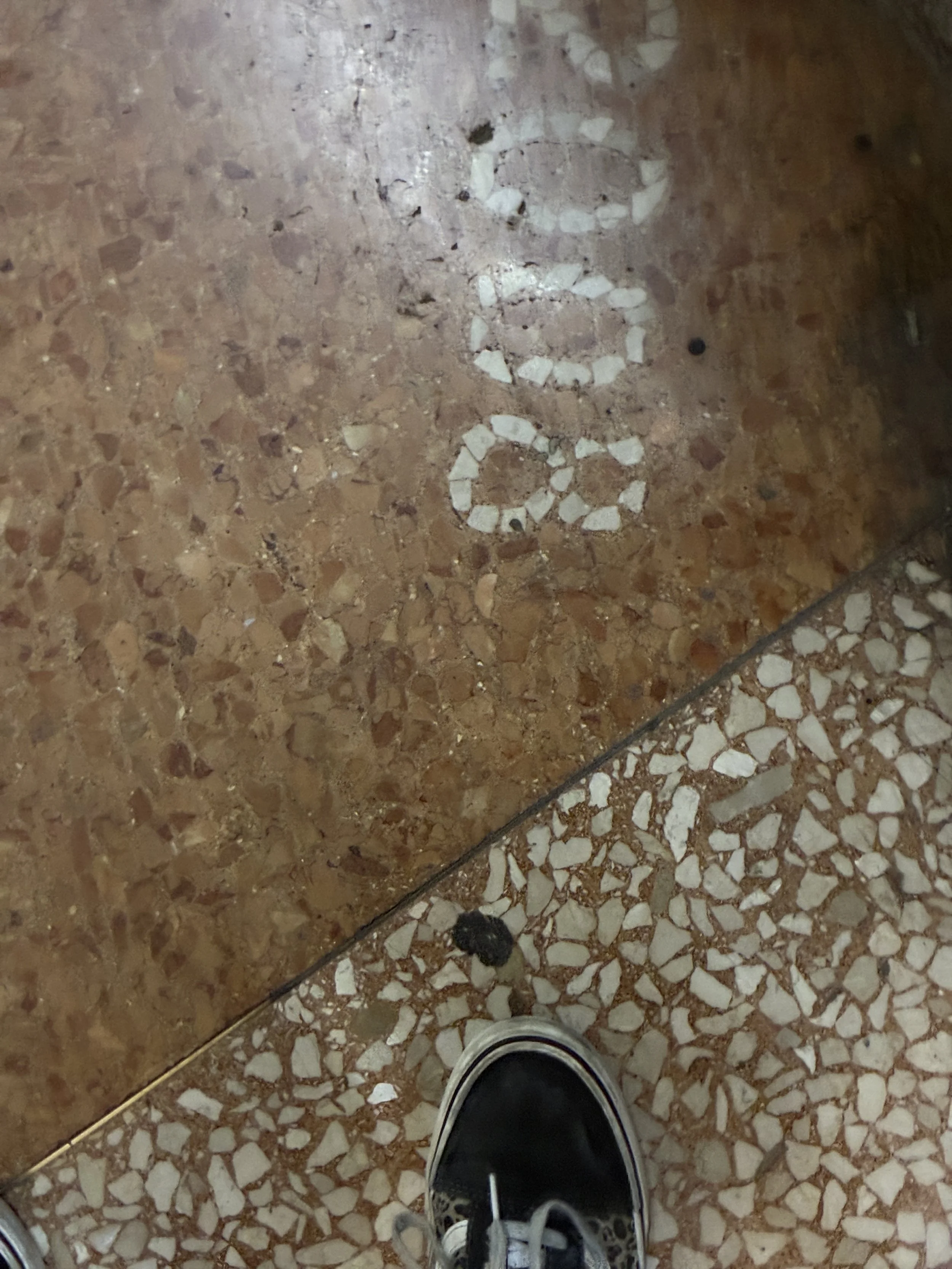 Close-up of a tiled floor with painted white initials 'c c c' on the surface and a person's sneaker at the bottom of the image.