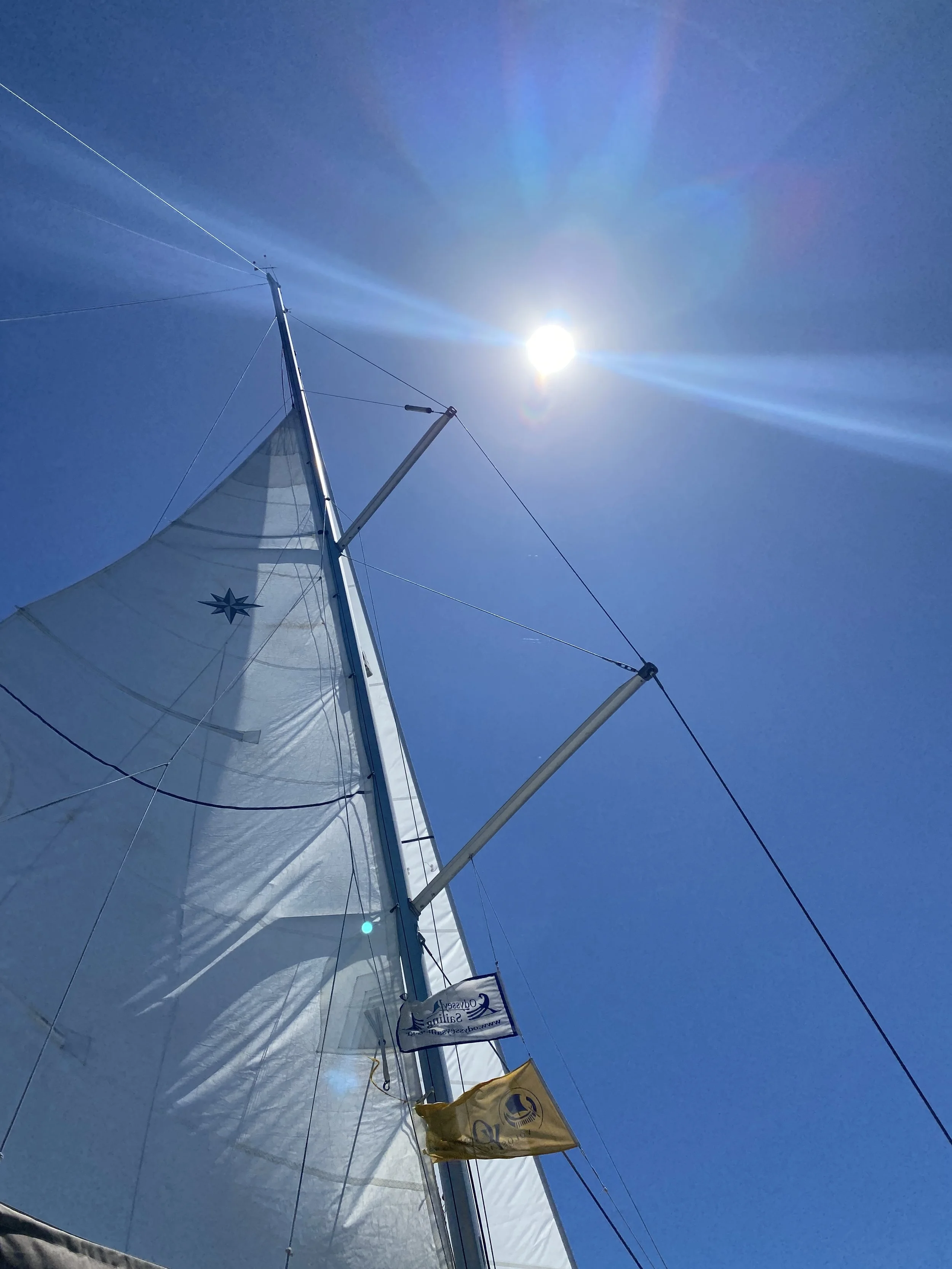 View of a sailboat mast and sail against a blue sky with the sun shining brightly.