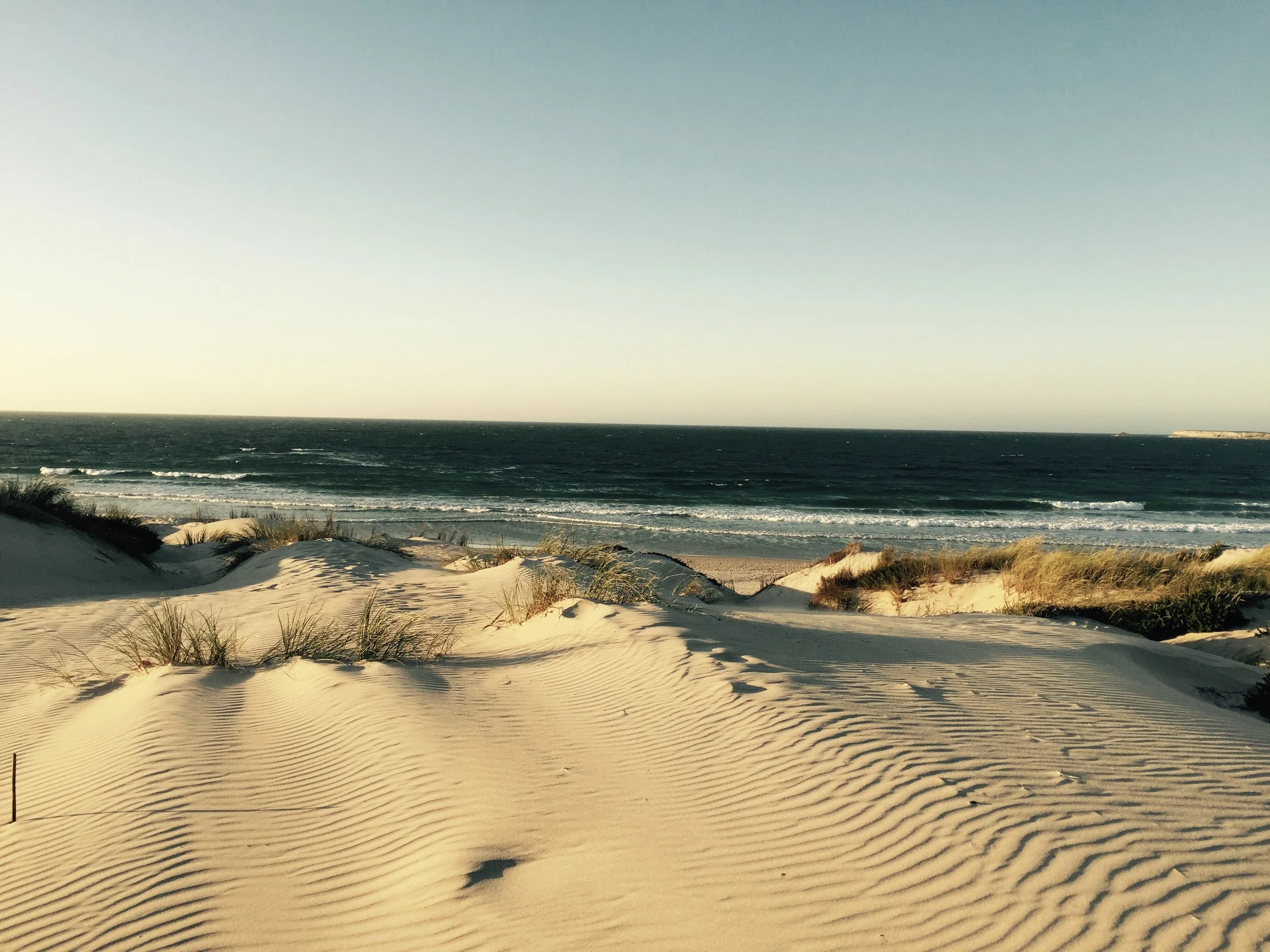 Sand dunes in the foreground with rippled patterns, the ocean with small waves in the background, and a clear sky with a gradient from pale yellow near the horizon to blue at the top.