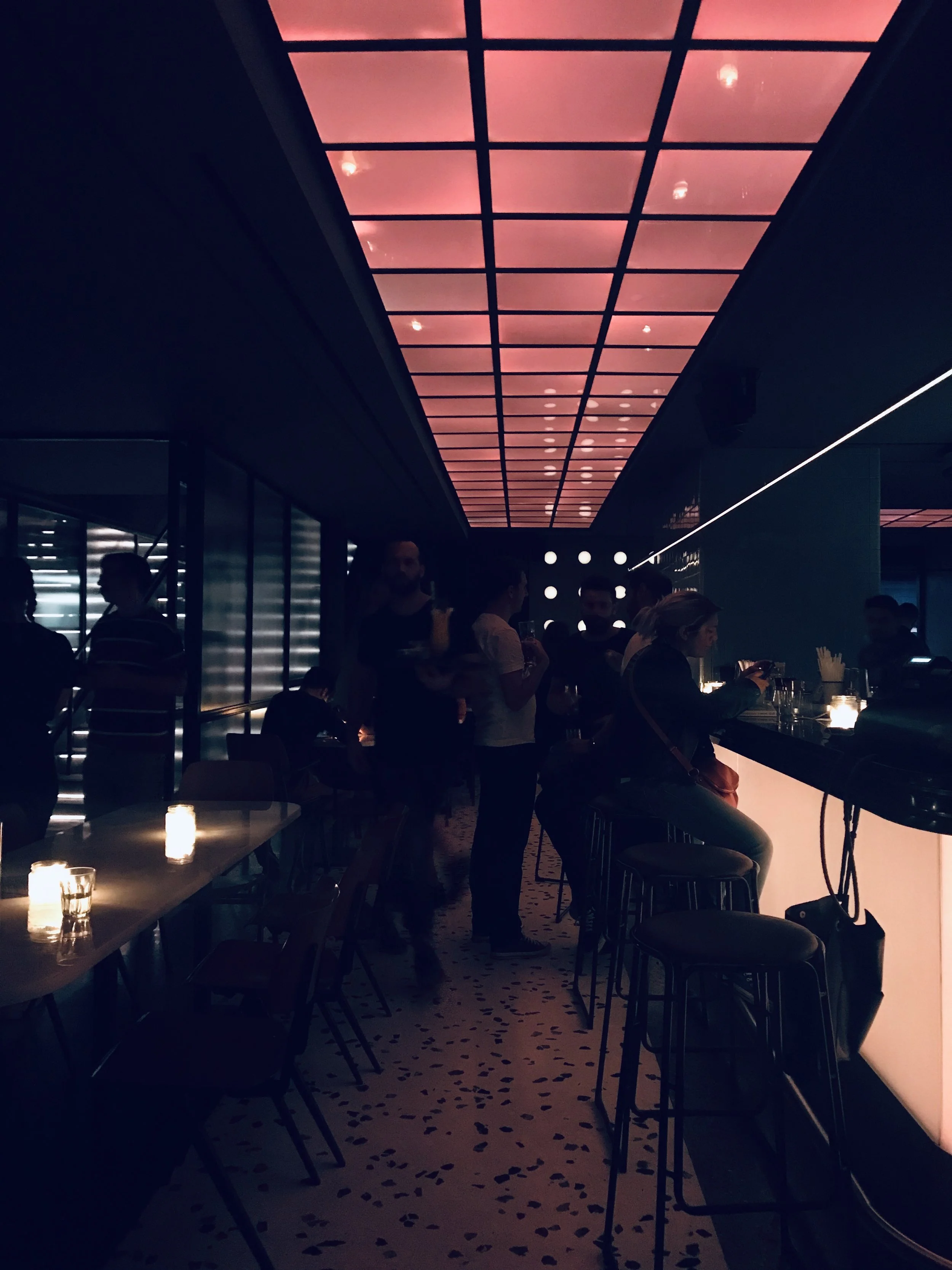 A dimly lit bar or lounge with a modern ceiling design emitting pinkish-orange light. People are gathered around the bar and sitting at tables, and there are candles on the tables providing additional lighting.