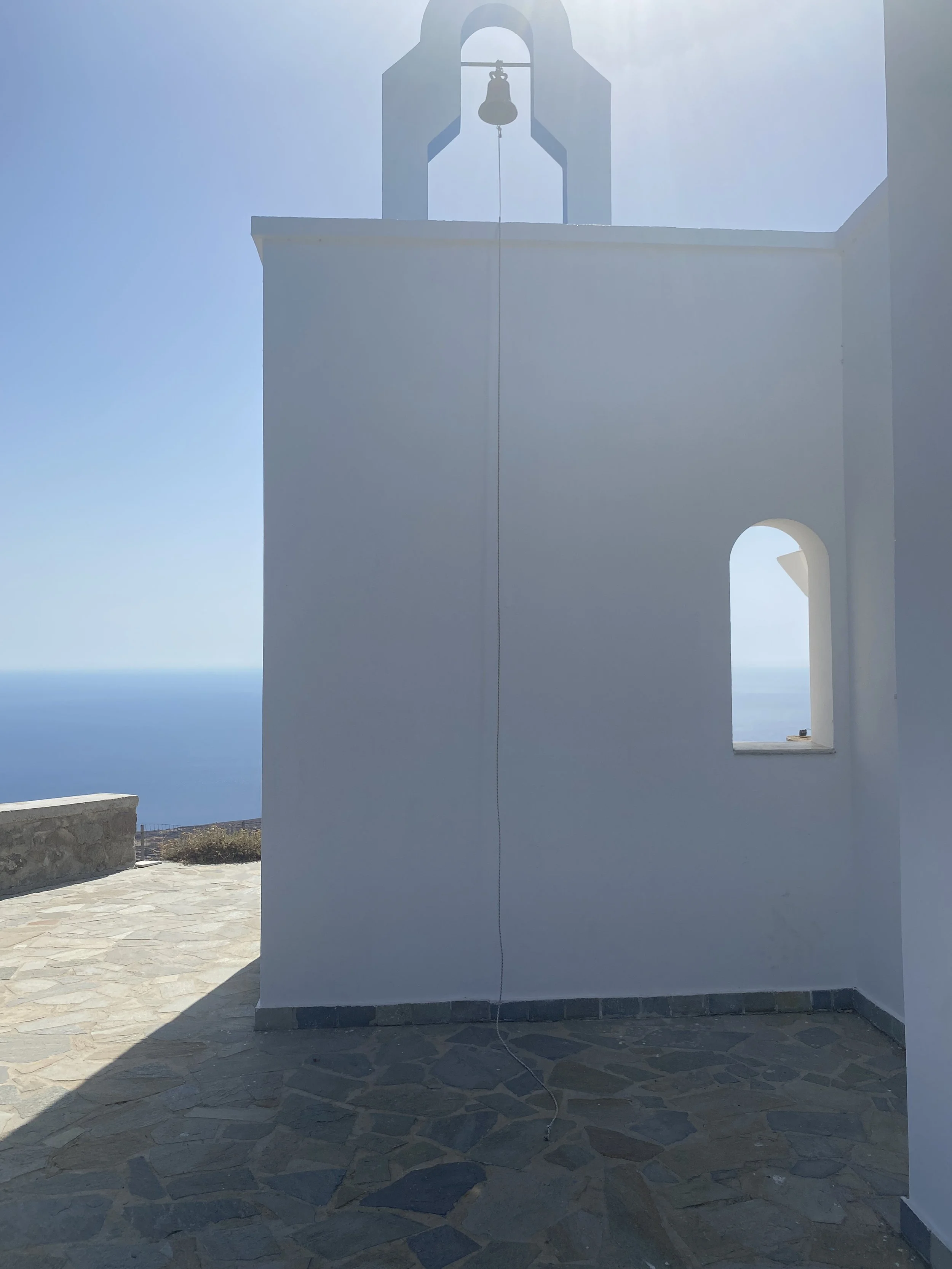 A white church tower with a bell at the top, seen against a clear blue sky with the sea in the background.
