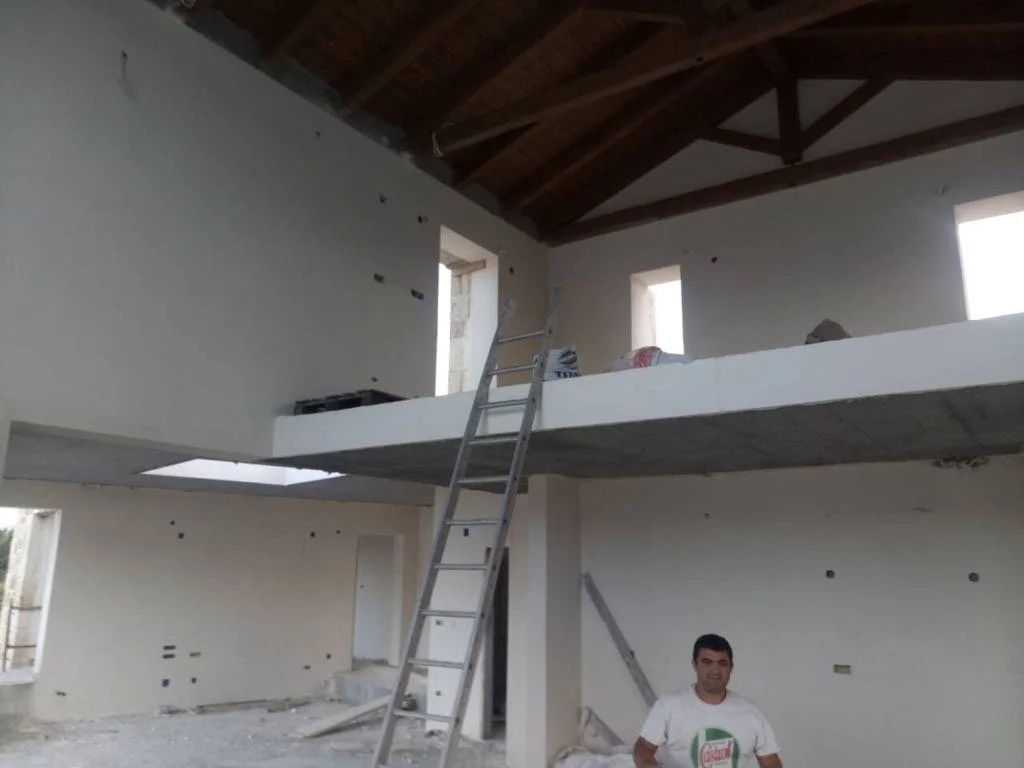 Interior view of a building under construction with a man smiling in the foreground. There are windows, a staircase, and a partially completed upper floor.