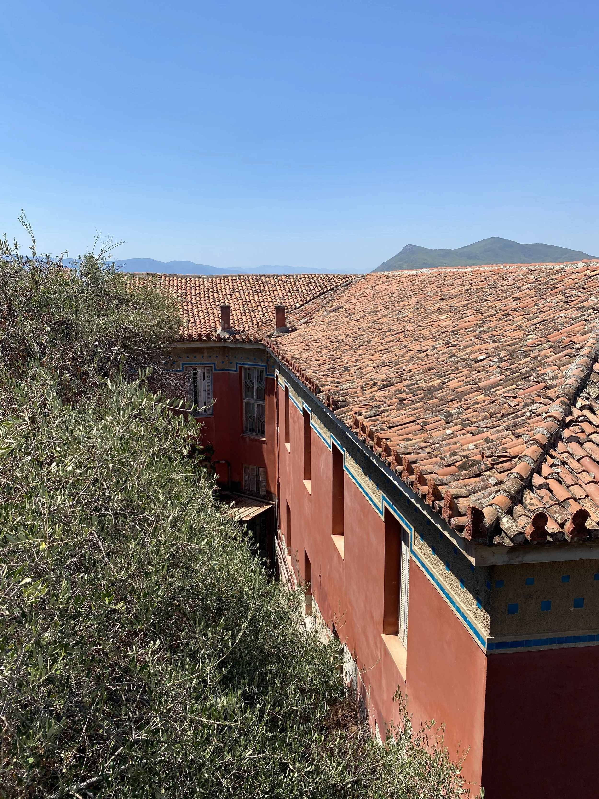 Building with pink walls and a red tiled roof, surrounded by vegetation, with mountains in the background under a clear blue sky.
