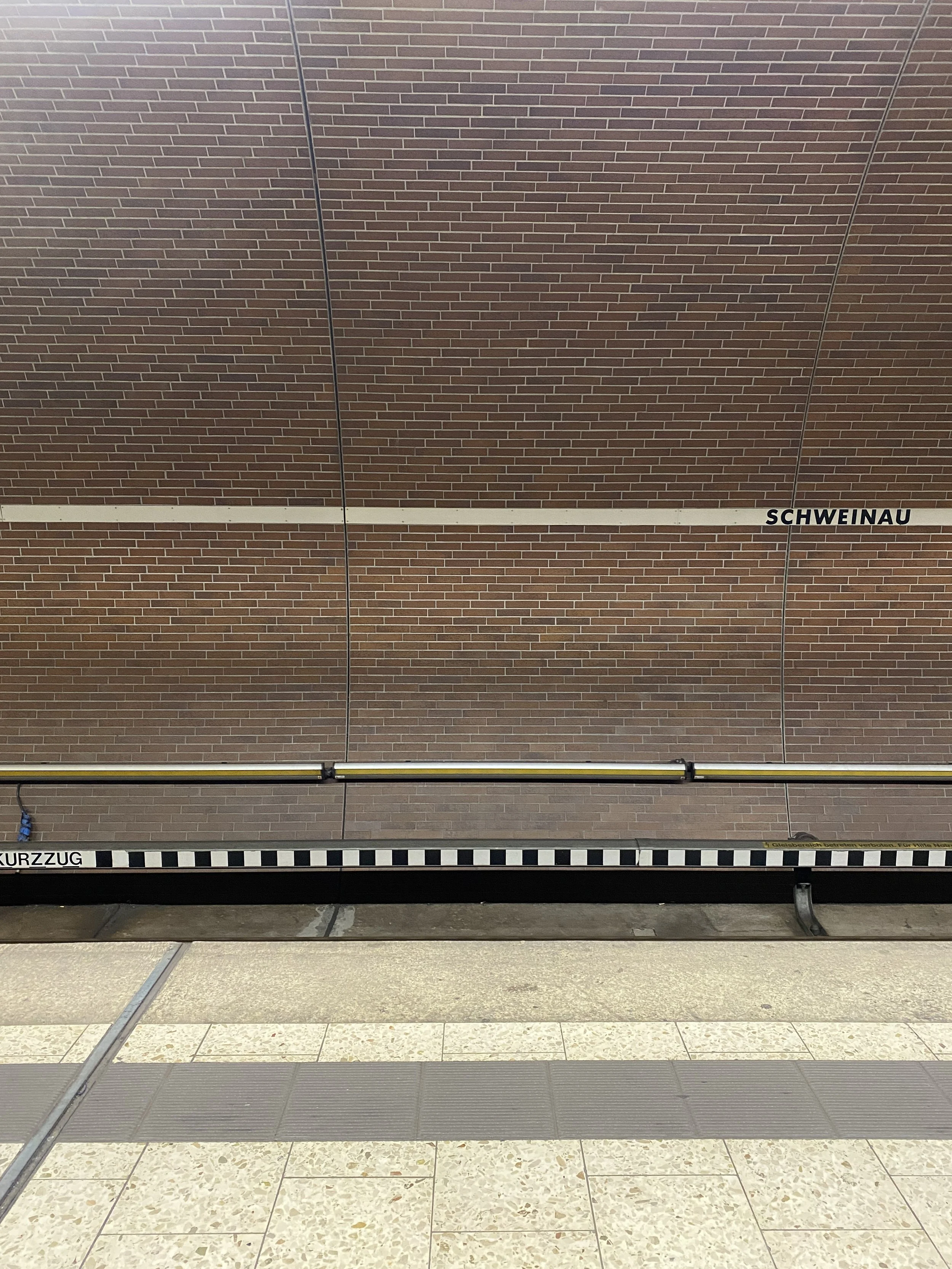 A brick wall with two black metal signs reading 'SCHWEINAU' and 'KURZZUG' and a yellow and black striped barrier or track in front of the wall.