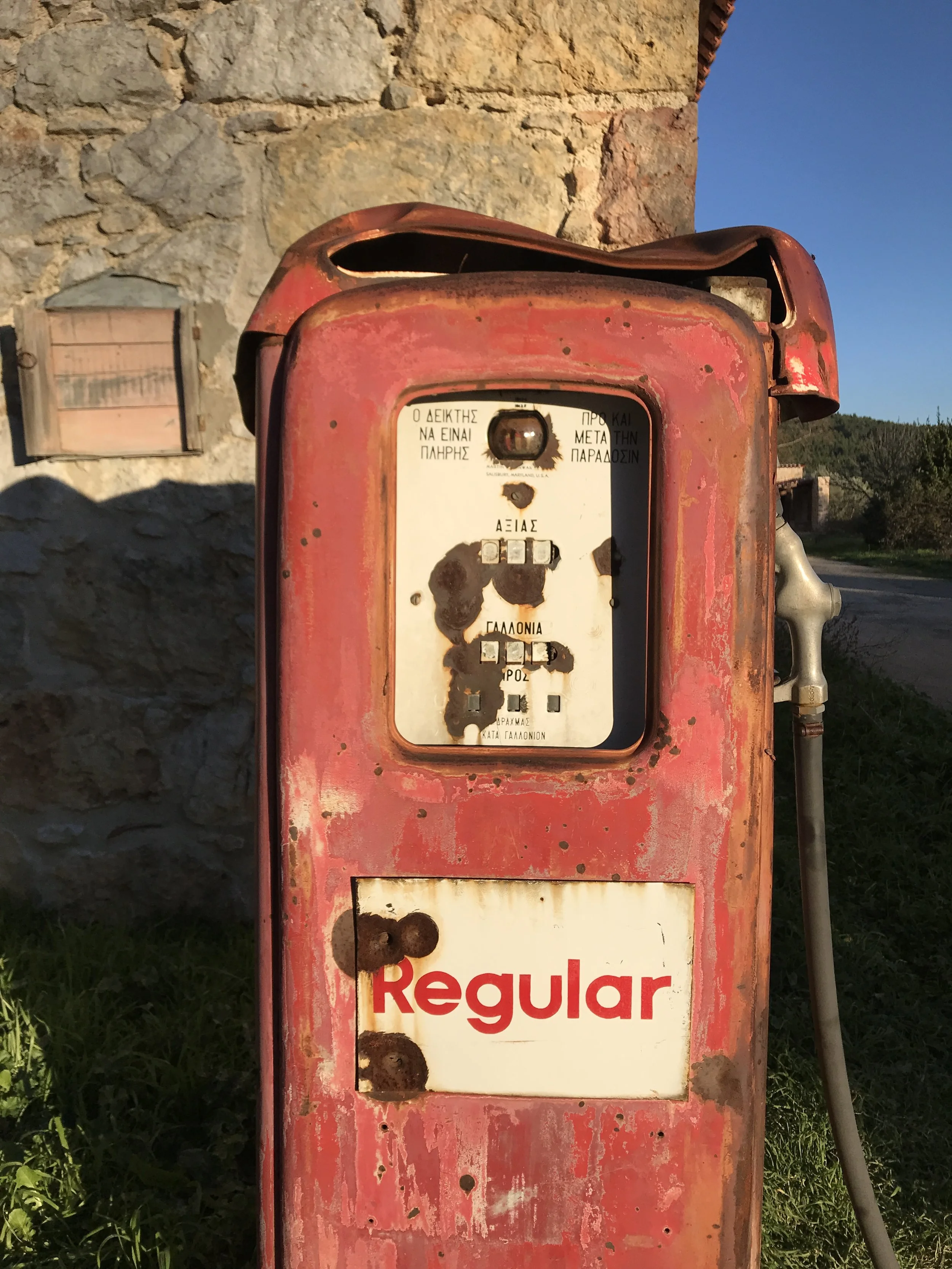 An old, rusty red gas pump with a partially missing faceplate, showing worn-out labels and a message in Greek. The patina and rust indicate age and exposure to the elements. In the background, there is a stone wall, a small window, and clear blue sky