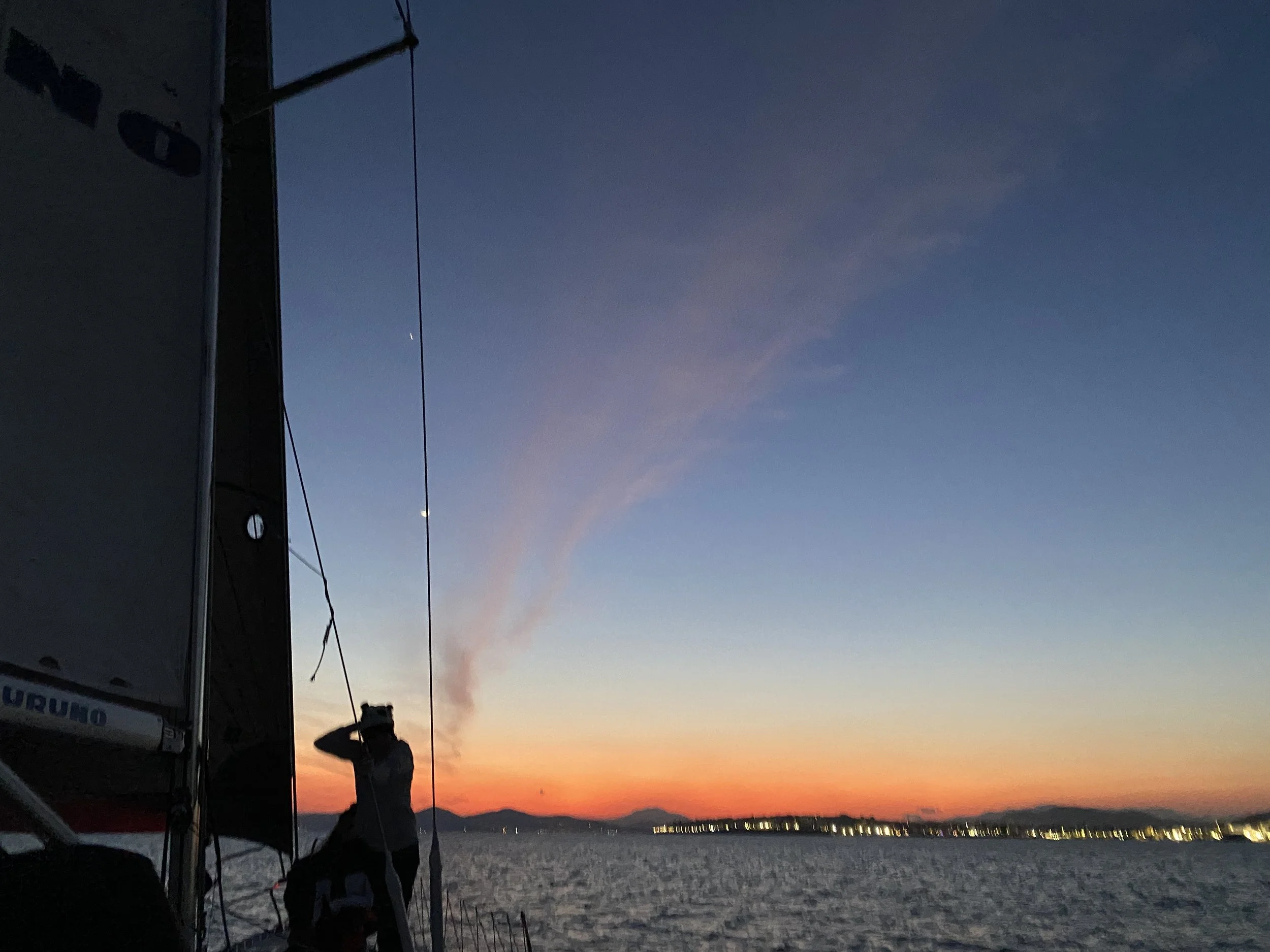 A person standing on a boat at sunset with a colorful sky and distant city lights