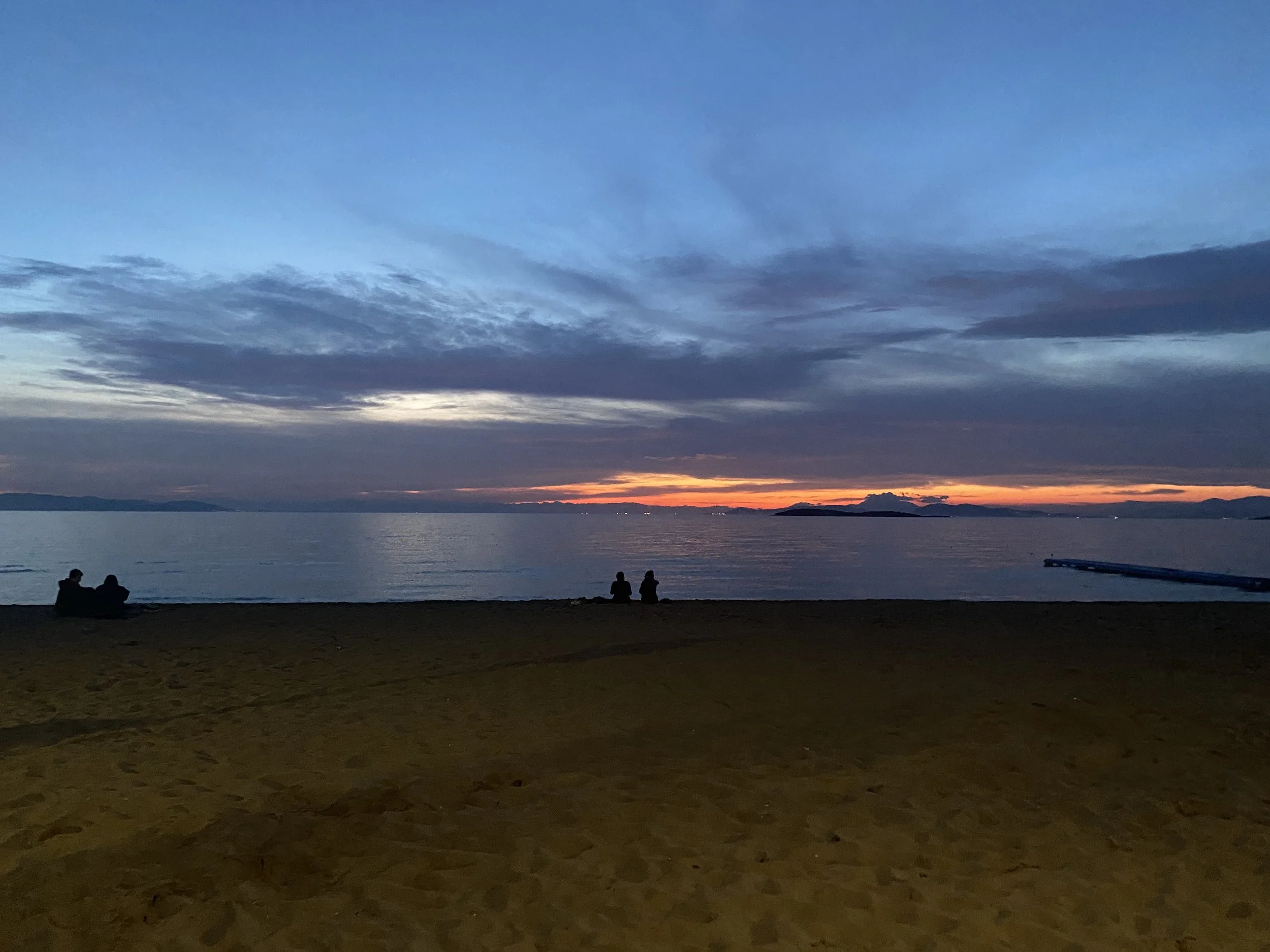 A sunset over a body of water with a few silhouetted people sitting on the sandy beach in the foreground, and mountains in the distance.