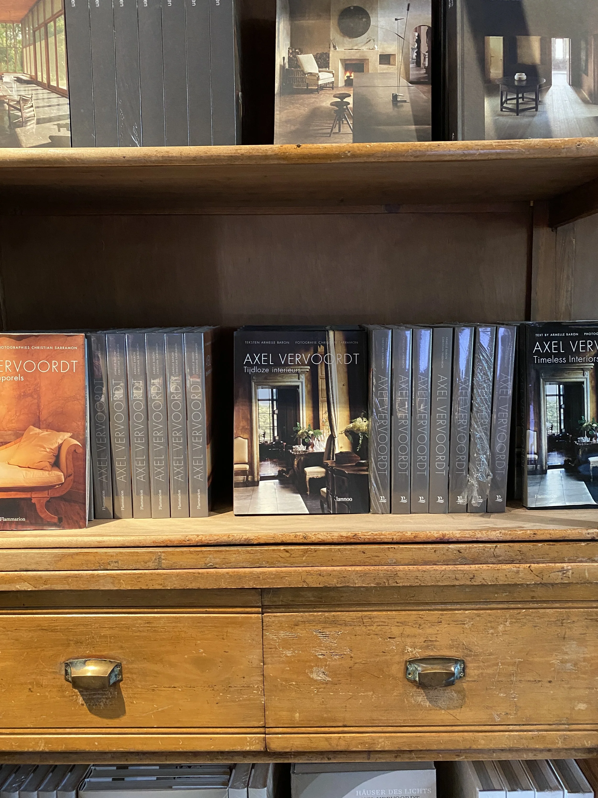 Wooden bookshelf containing books, with a view through a window in the background showing a cozy interior with a fireplace, table, and chairs.
