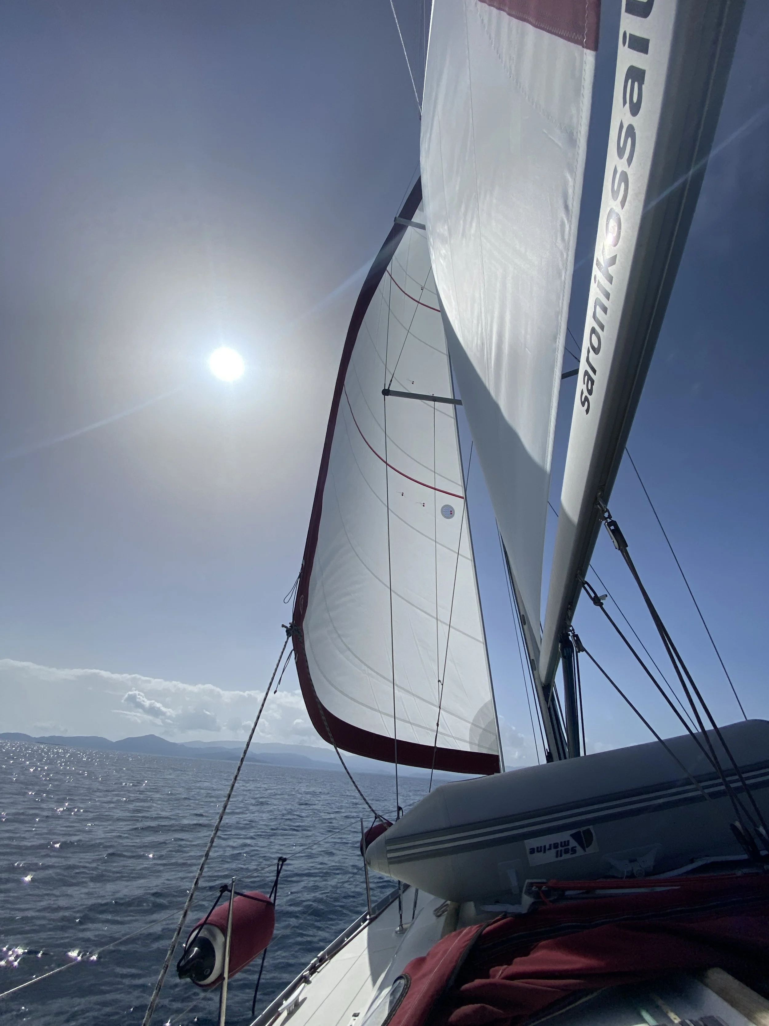 View from a sailboat at sea during daytime, showing sails and the horizon with distant land and mountains under a clear sky with the sun.