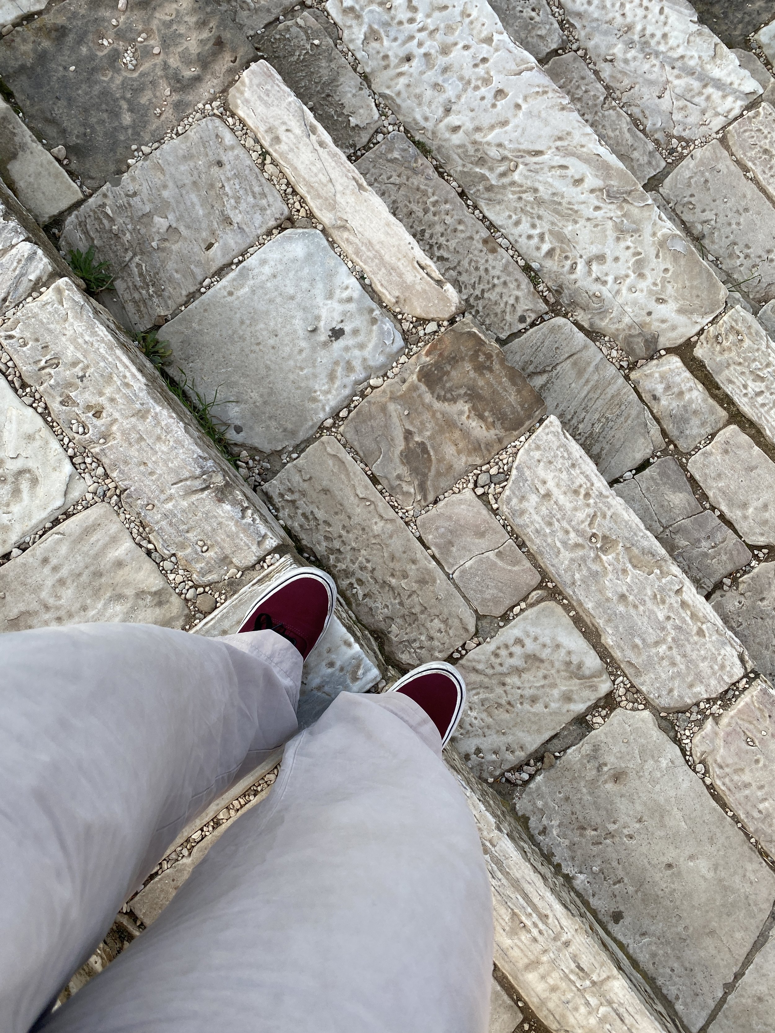 A person's legs and feet in red sneakers and white pants standing on a cobblestone street.
