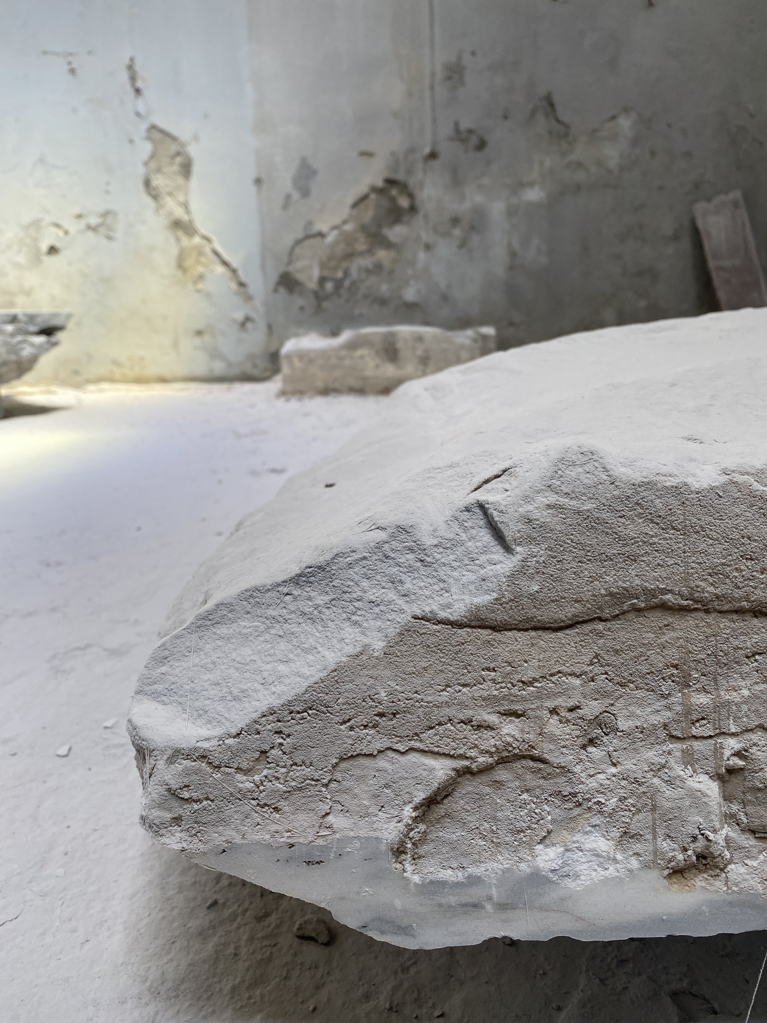Close-up of a textured, white archaeological artifact or sculpture with faint carved lines, in front of a weathered stone wall.