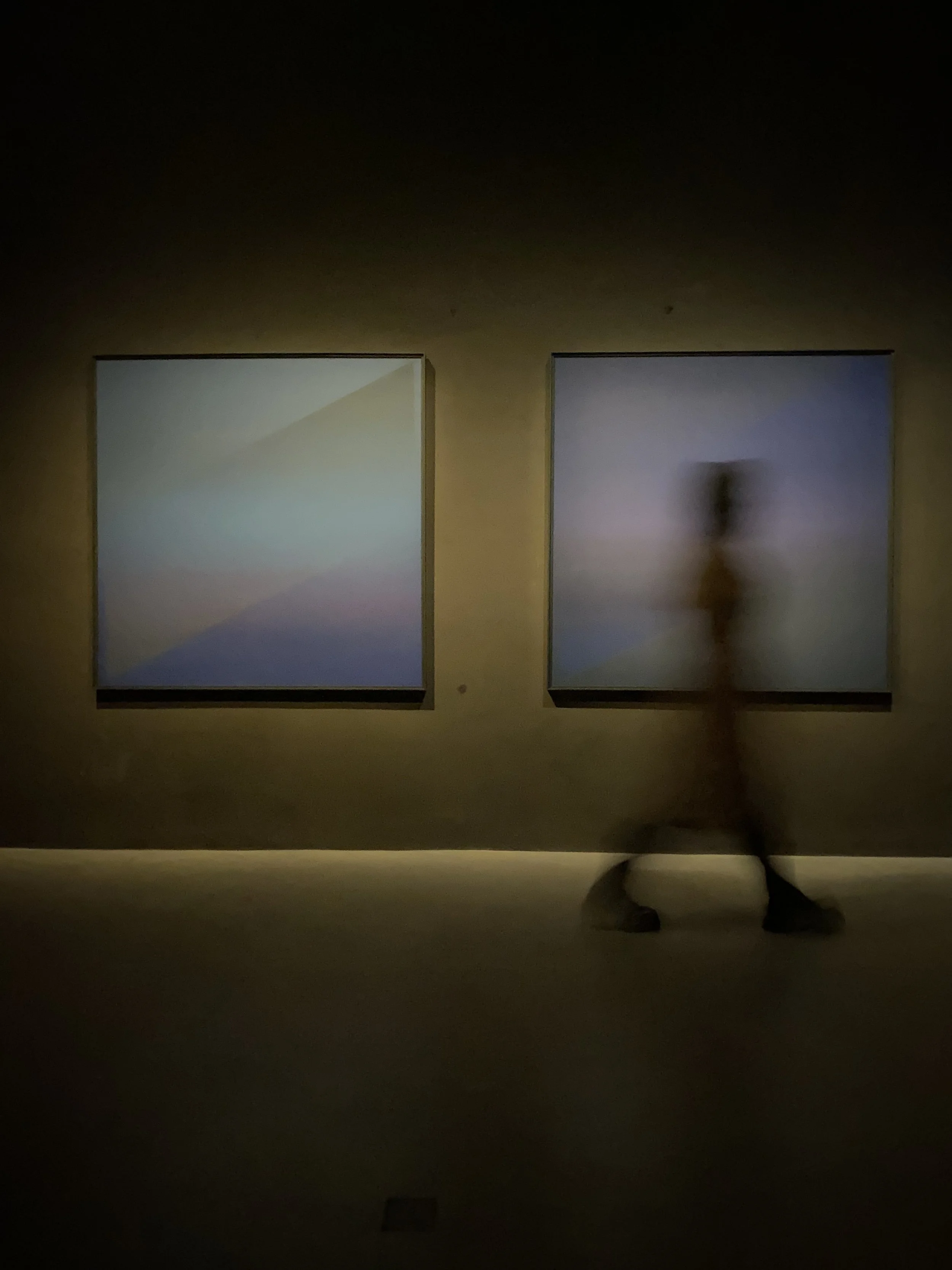 A blurred silhouette of a person inside an art gallery, standing in front of two abstract paintings with dark frames, featuring gradient colors and soft light.