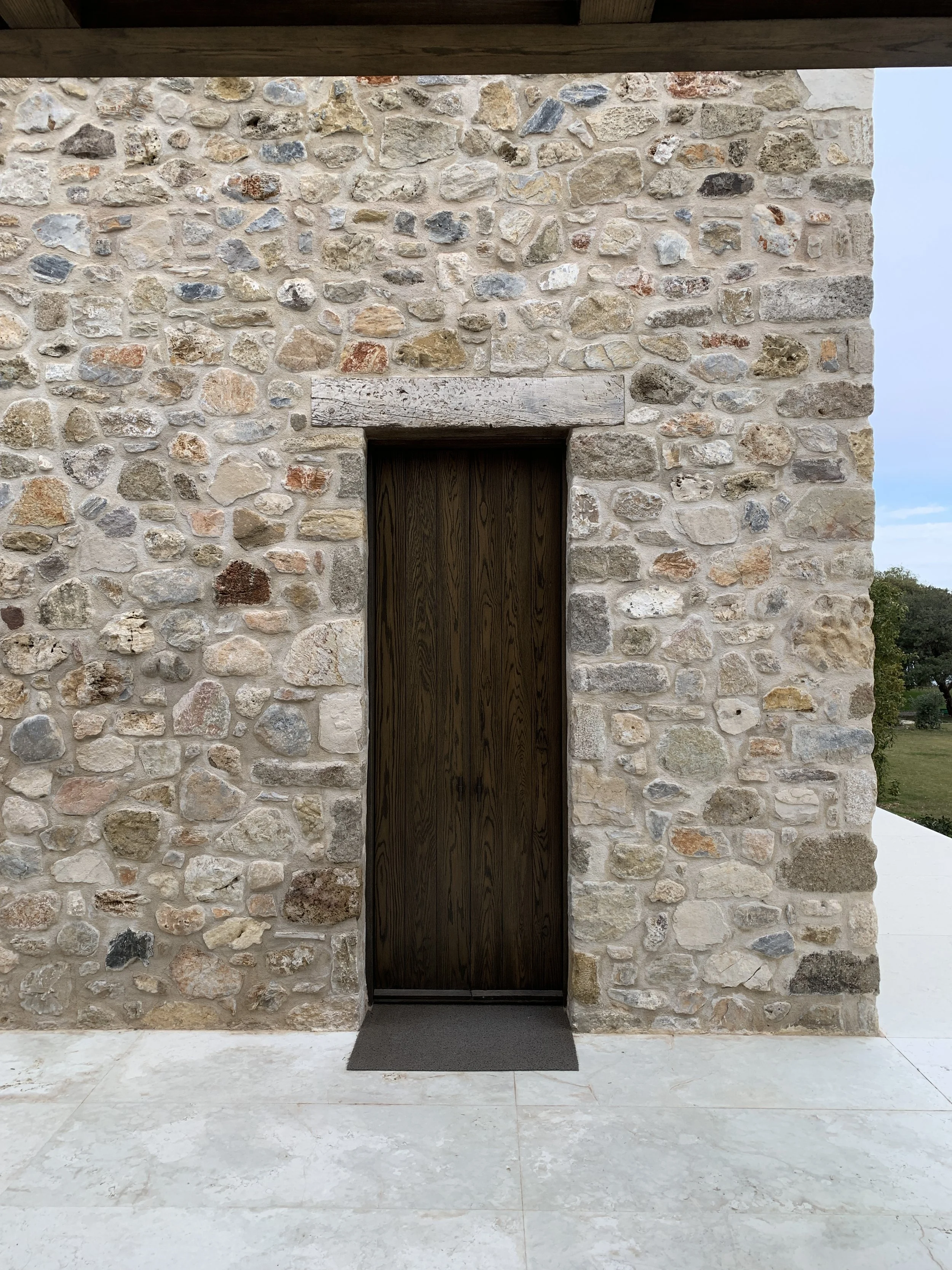 Stone wall exterior with a dark wooden door and a small gray mat in front of it, under a covered porch area with wooden ceiling.