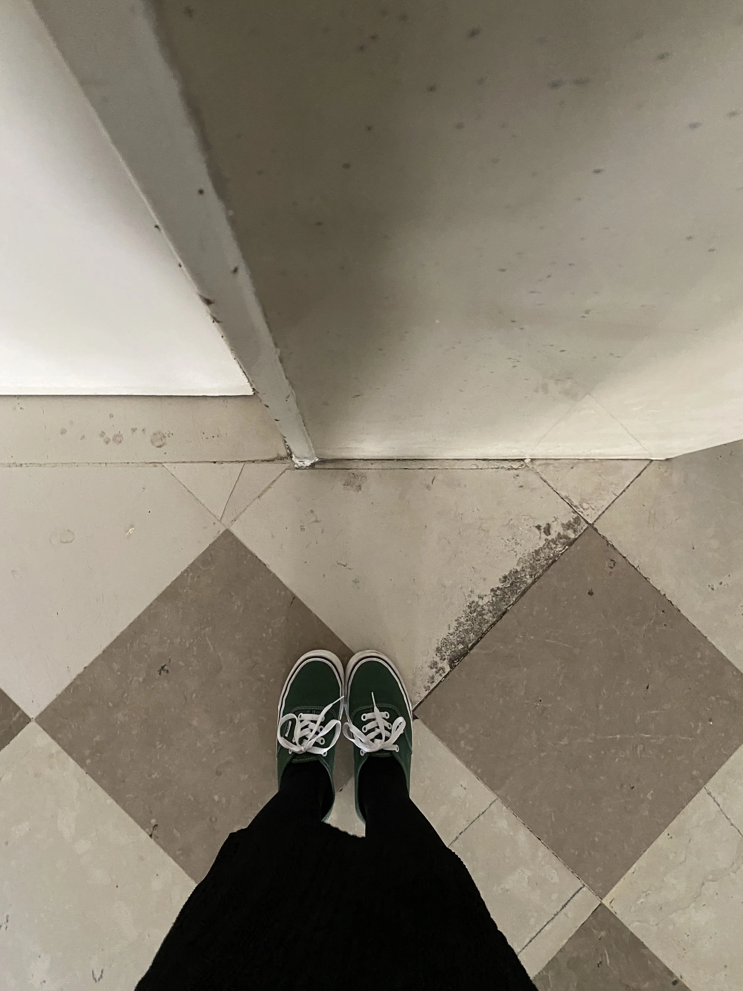 Photo showing a person standing on black and white checkerboard patterned tile floor, wearing black pants and green sneakers with white laces, near a white door and a wall with some dirt and grime.