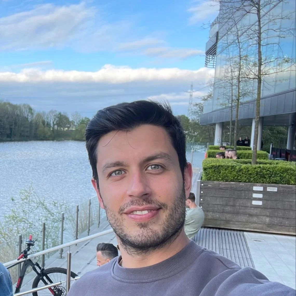 Male with tan skin tone and black hair and green eyes. Wearing a grey sweater. Standing outside with river in the background.